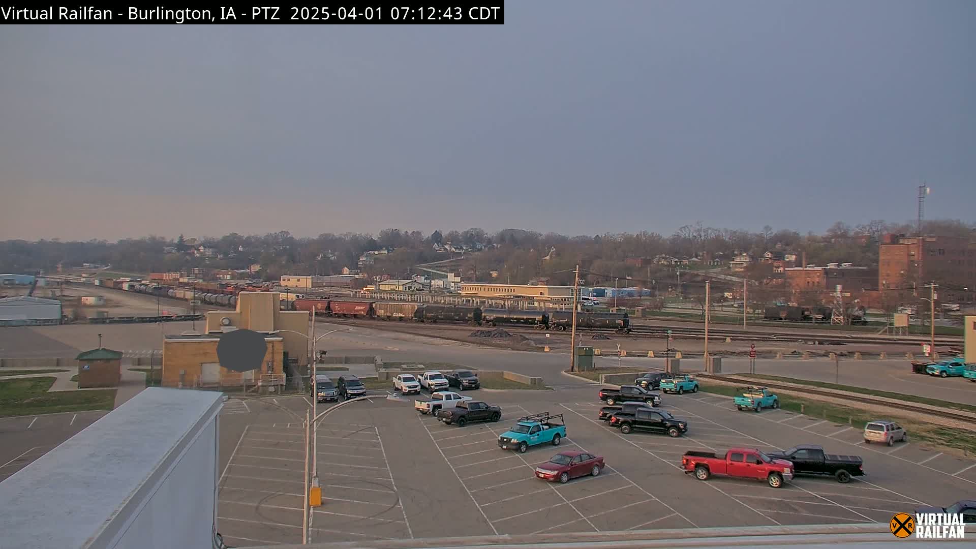A mostly clear, twilight sky overlooks a train yard with several railcars and a parking lot containing numerous vehicles.