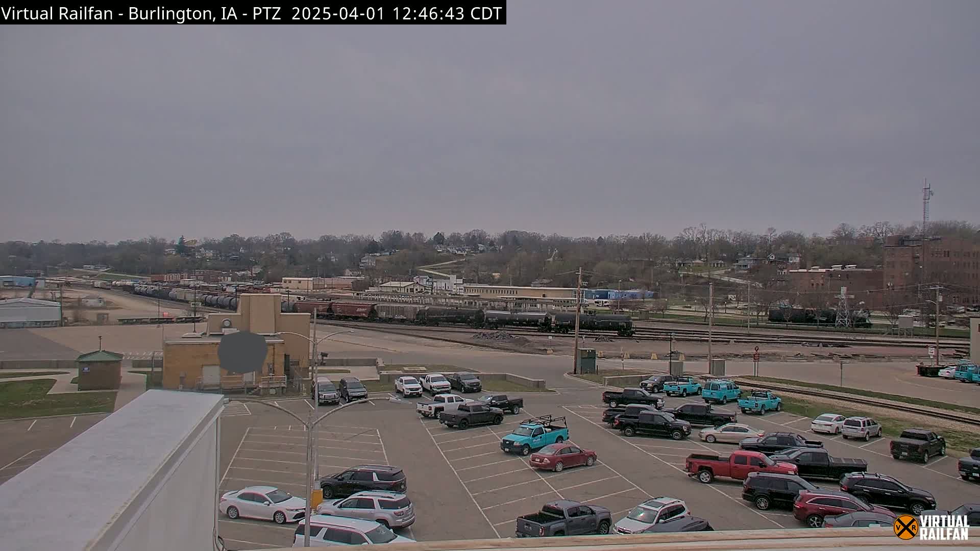 A large parking lot filled with cars is situated near a train yard with numerous railcars under an overcast sky.