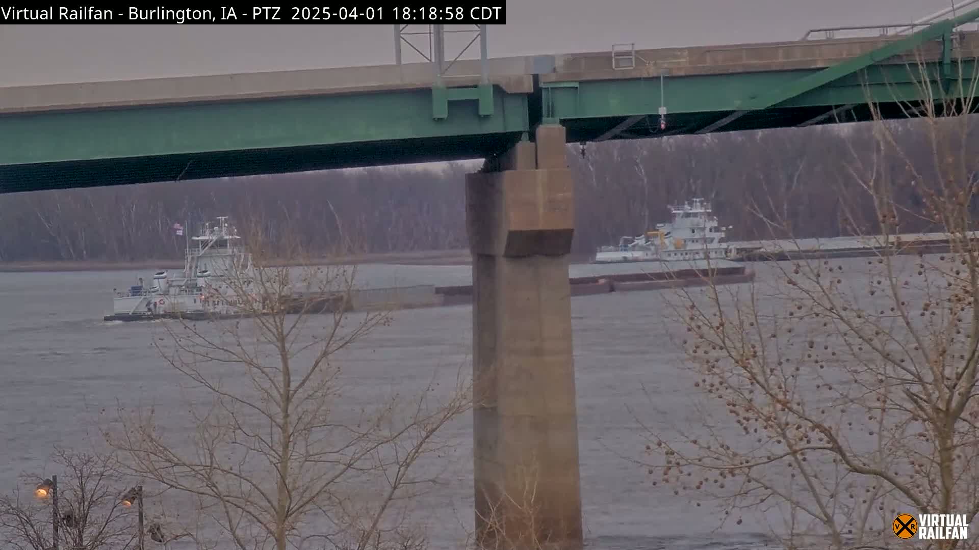 Two towboats pushing barges are on a river under an overcast sky, viewed from under a bridge.