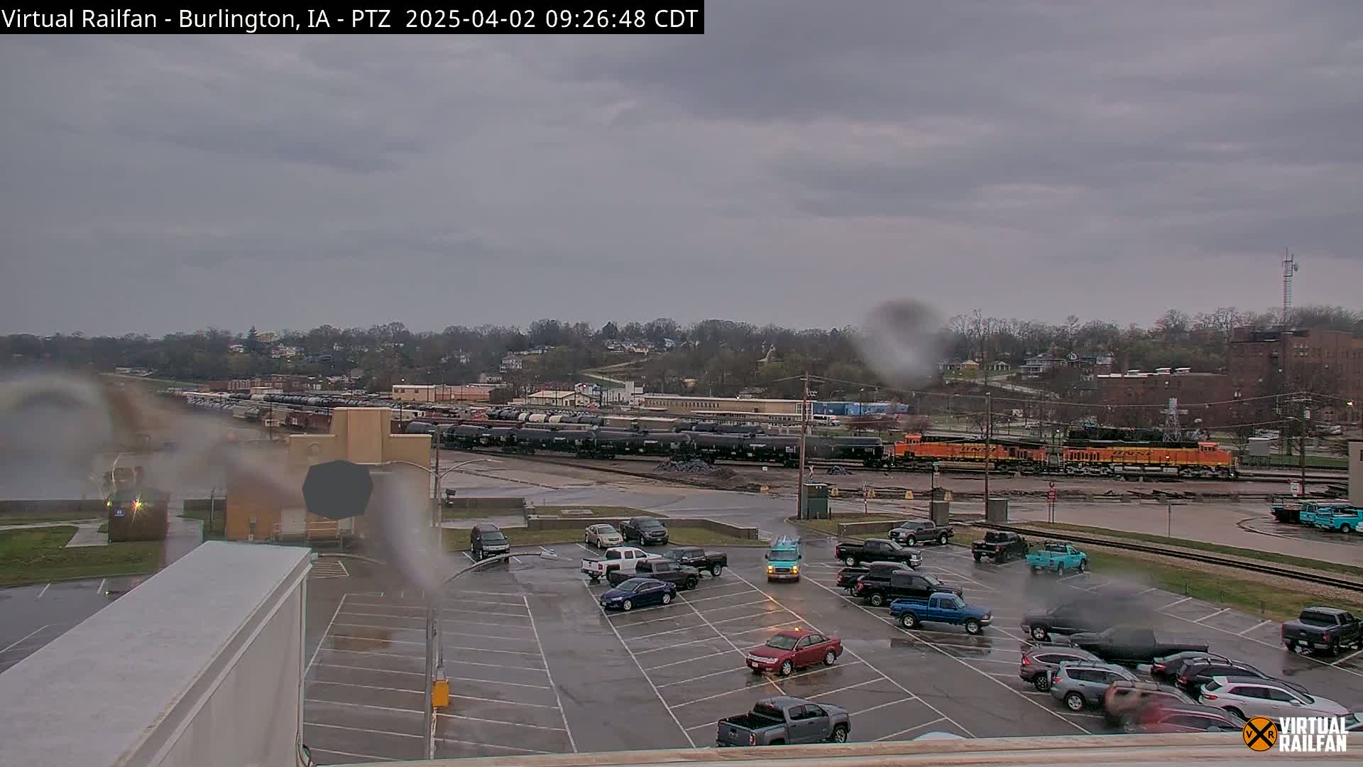 A rain-covered view shows a large parking lot with several cars, a train with orange locomotives in the background, and a tree-covered hillside beyond.