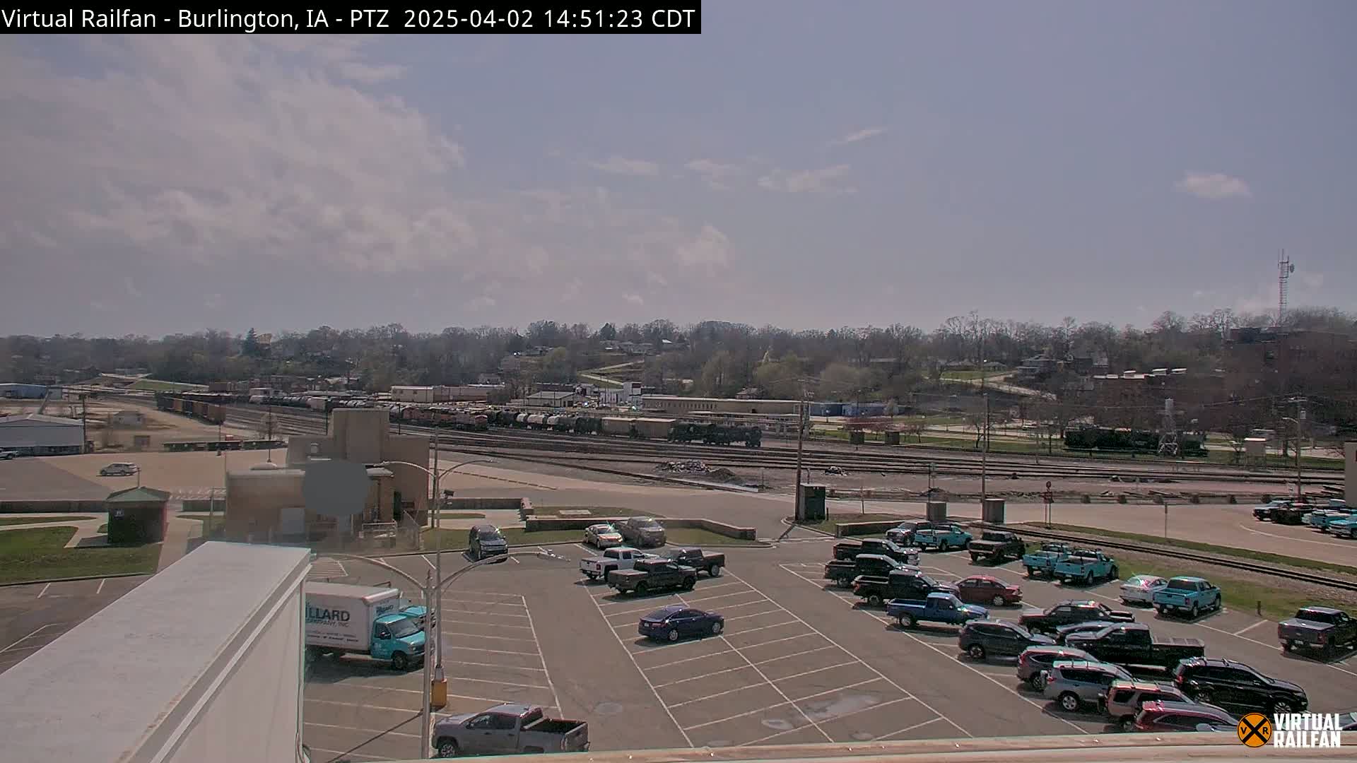 A mostly sunny day reveals a railway yard with numerous freight cars and a parking lot filled with various vehicles, all situated in front of a tree-lined hill.