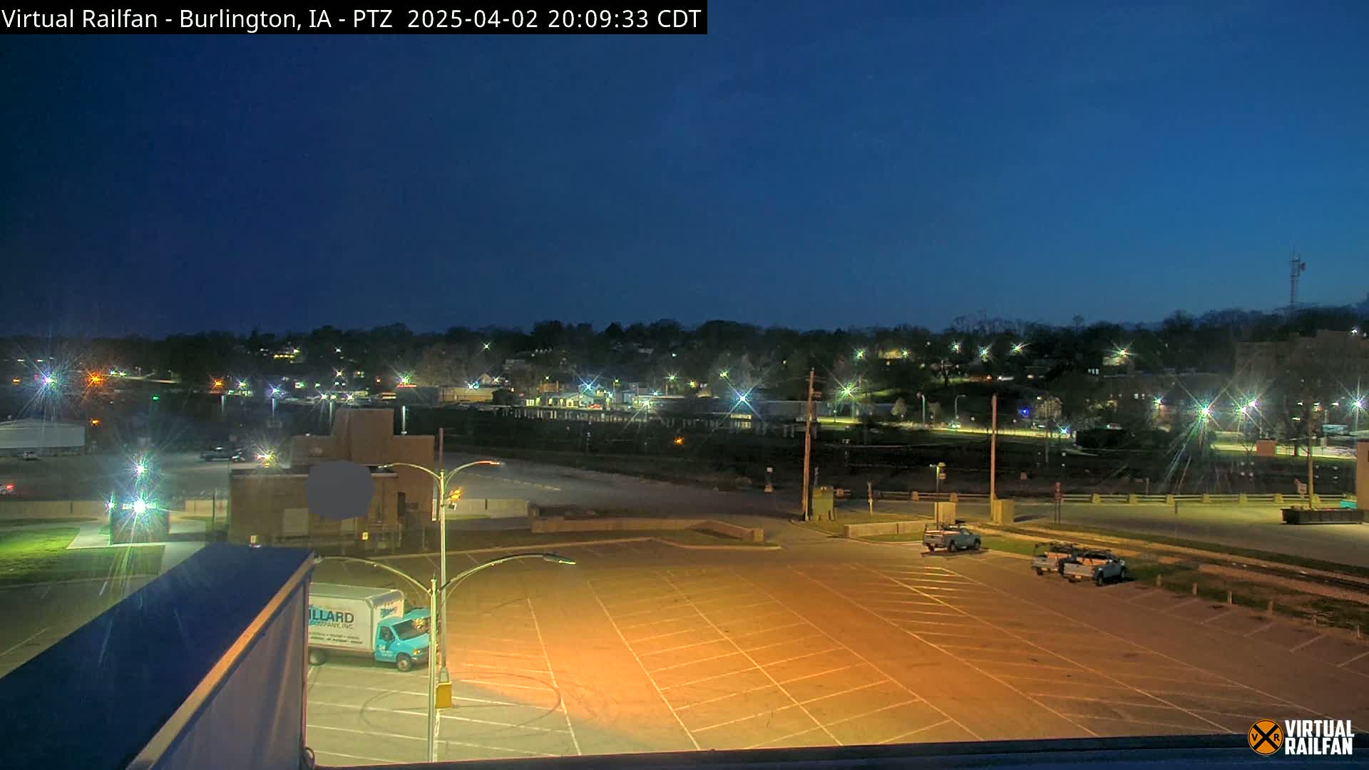 A mostly empty, illuminated parking lot at night, overlooking a town with streetlights and sparse nighttime traffic under a clear, dark blue sky.