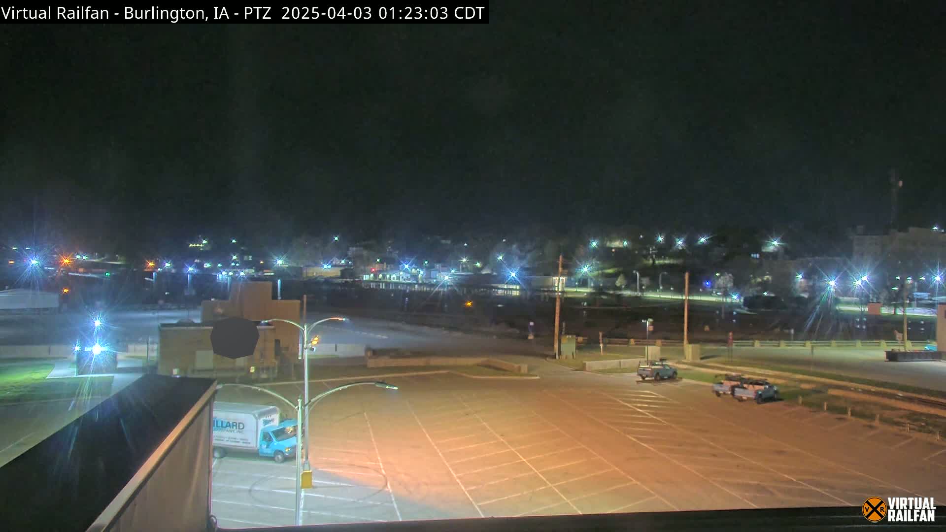 A mostly empty parking lot at night is shown, with a few vehicles and a small delivery truck present, and a town's lights visible in the background.