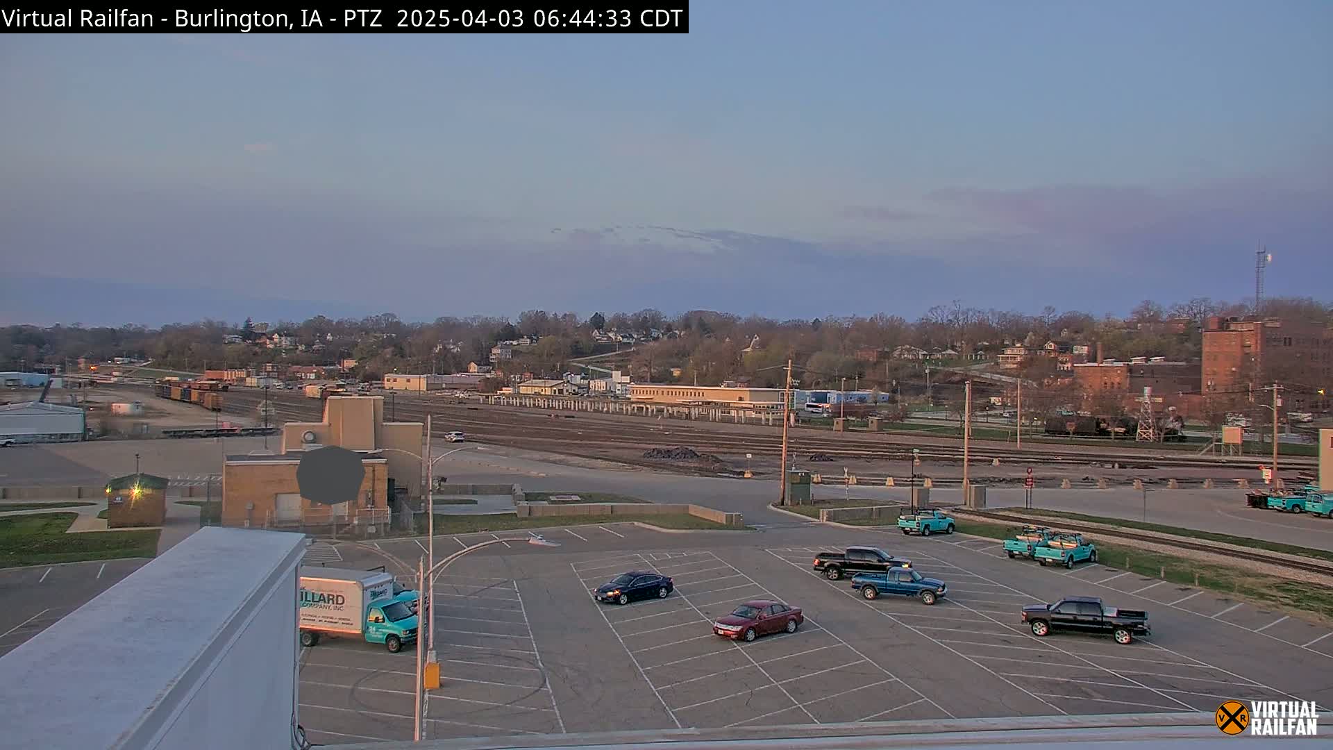 A mostly empty parking lot with several cars and trucks, situated near a railroad yard and industrial area under a twilight sky.