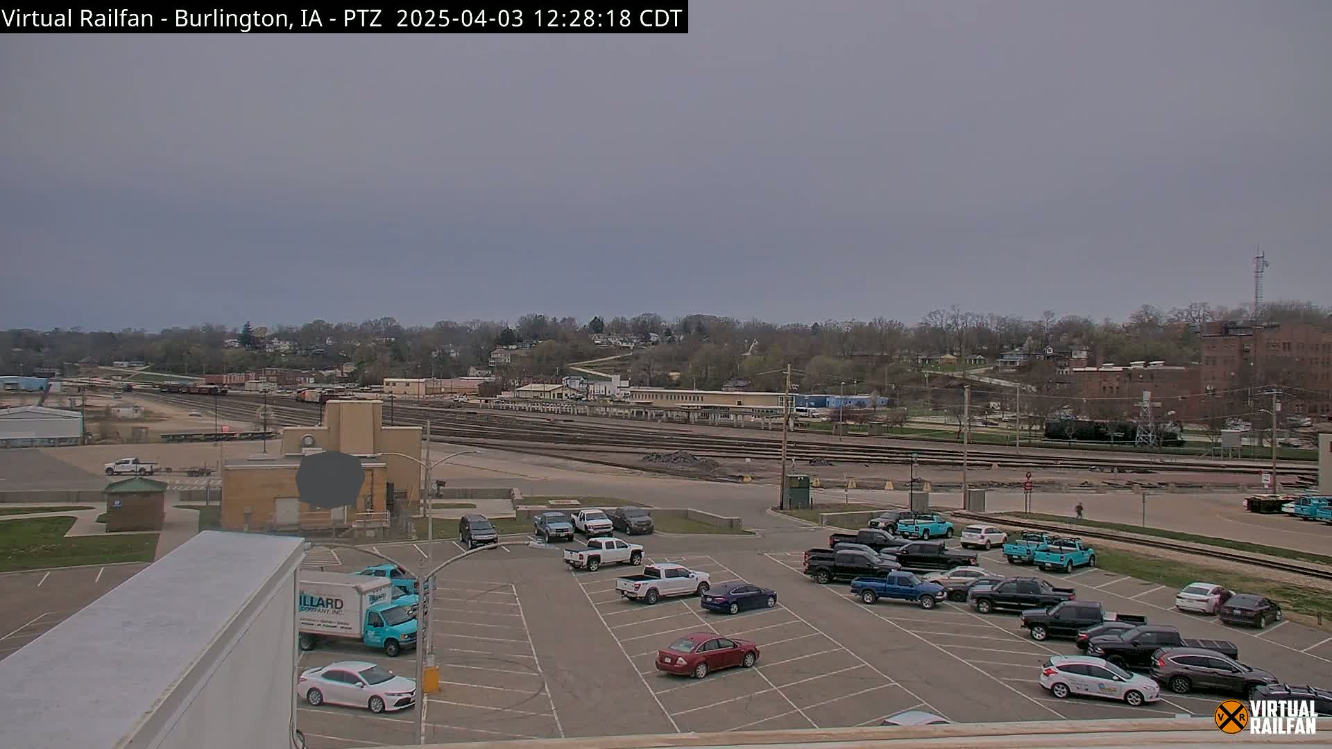 A mostly cloudy day reveals a large railroad yard with numerous train cars and a parking lot filled with cars and trucks.