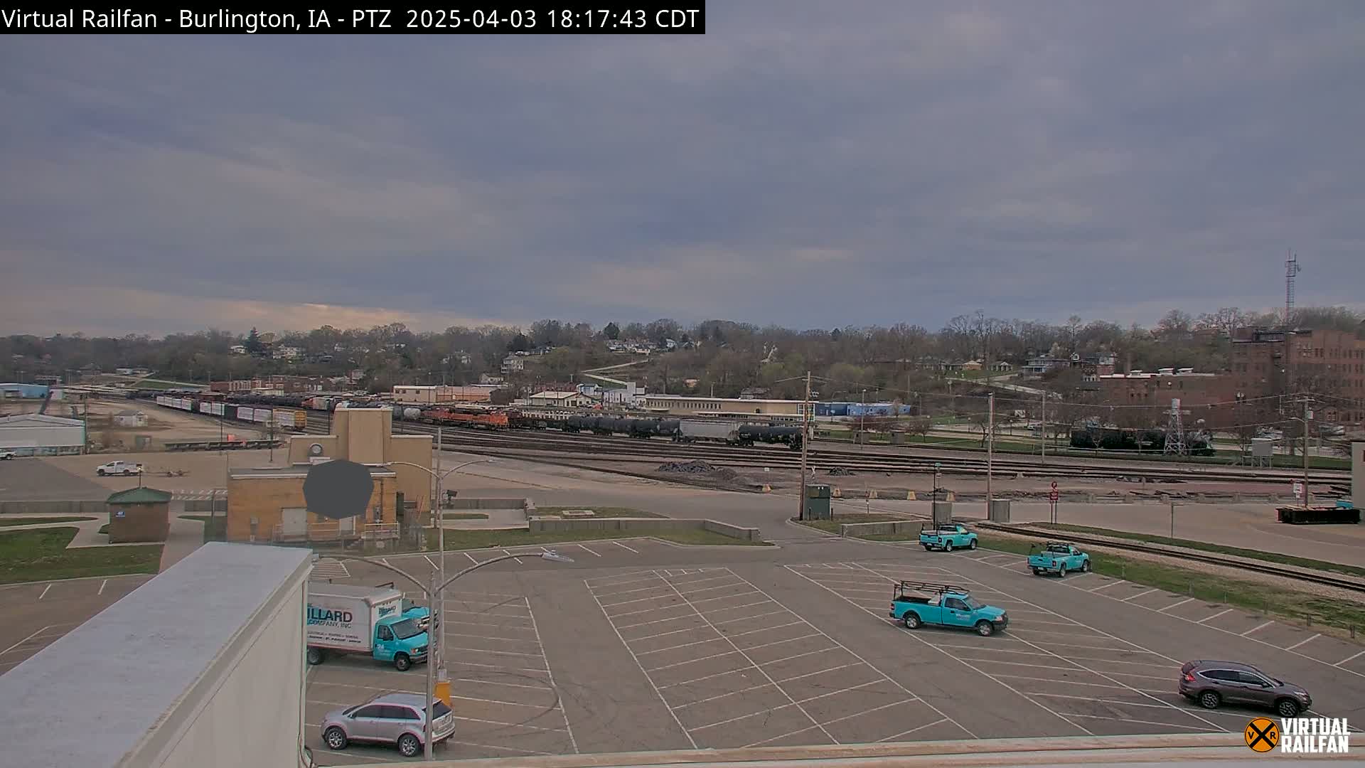 An overcast sky looms over a rail yard with several freight cars, a large empty parking lot with a few teal pickup trucks and cars, and a town in the background.