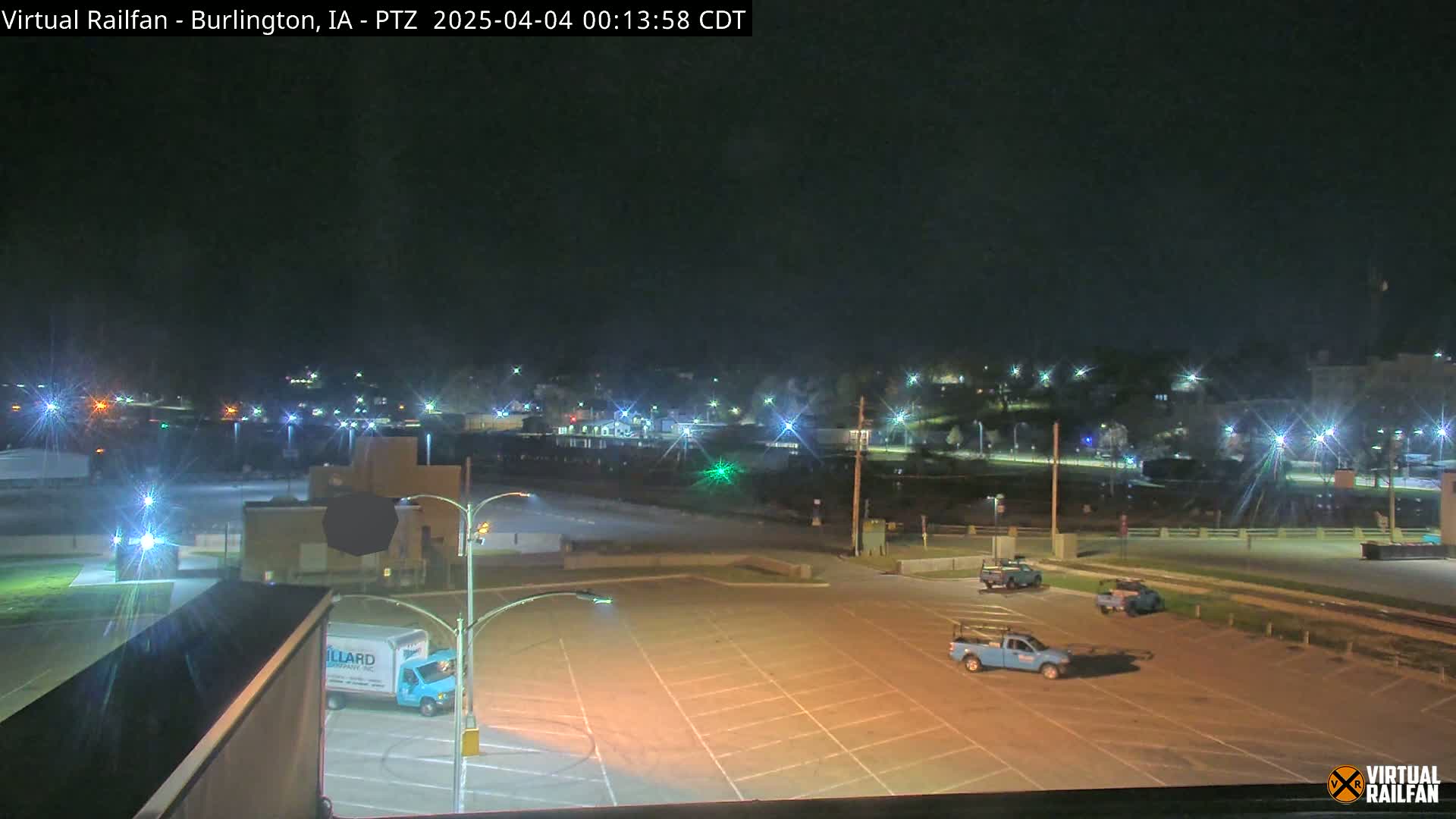 A nighttime view of a mostly empty parking lot with a few vehicles, illuminated by streetlights, overlooks a dimly lit town in the distance.