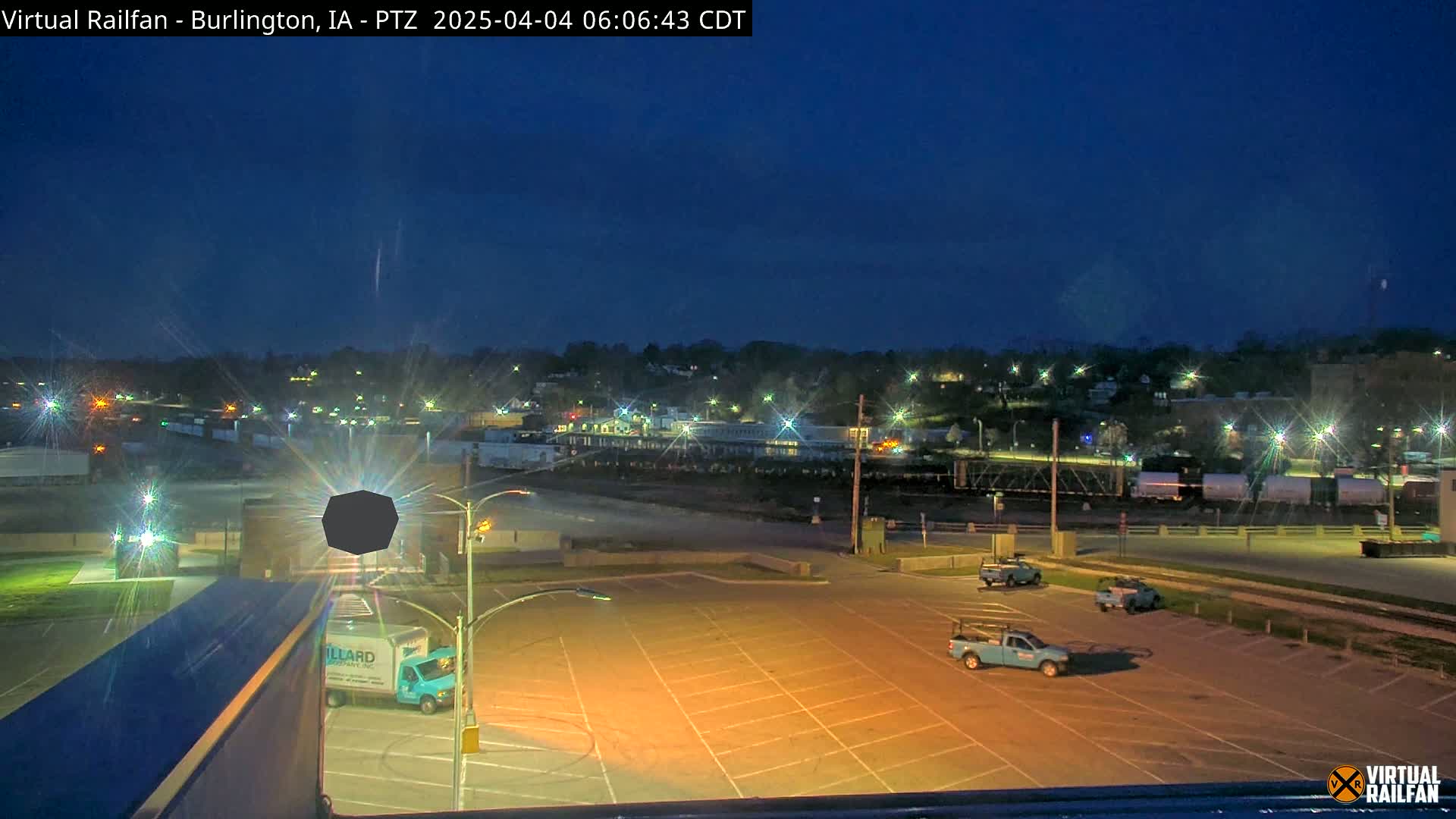 A nighttime view of a mostly empty parking lot with several trucks, train cars in the background, and a town faintly visible under a dark blue twilight sky.