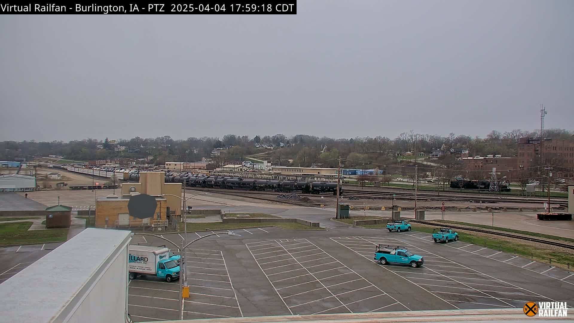 A large empty parking lot with several teal pickup trucks is adjacent to a train yard containing numerous railcars under an overcast sky.
