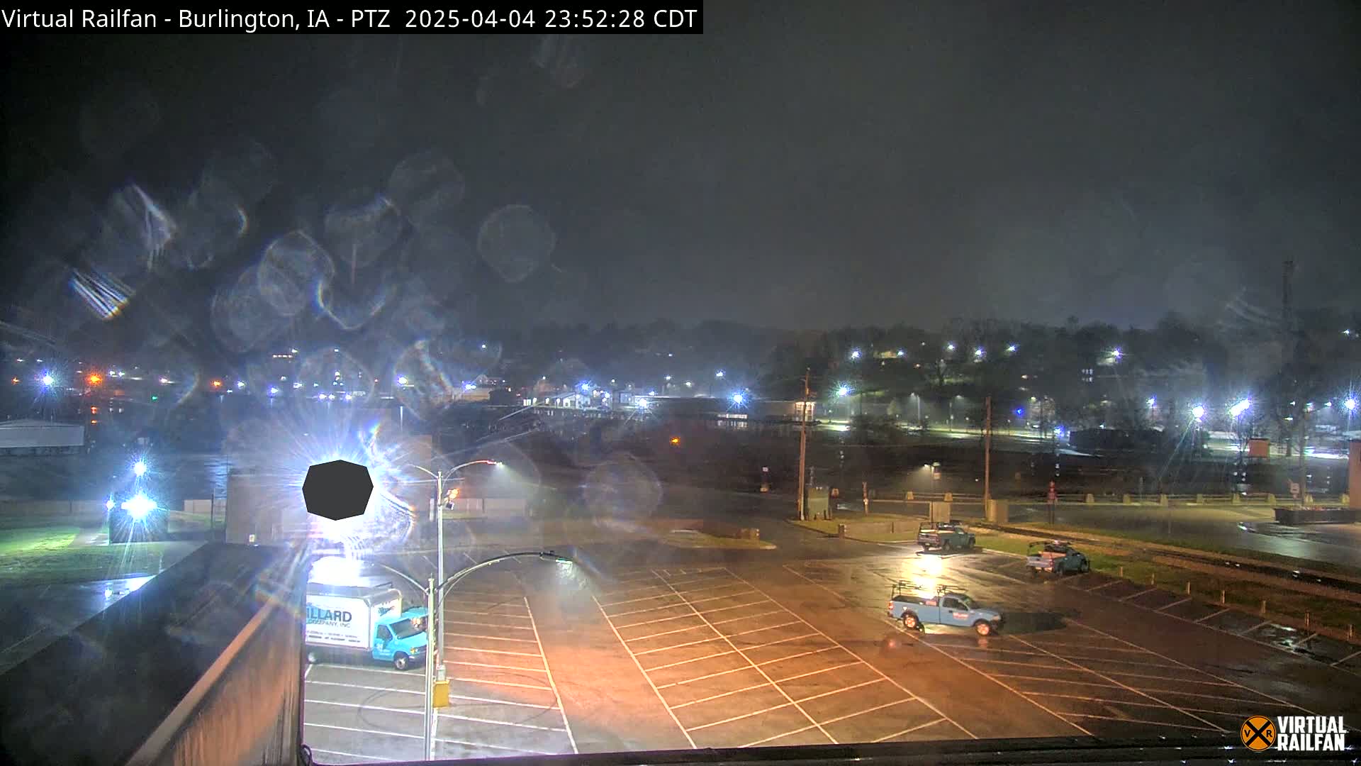 A rain-slicked parking lot at night, with a few vehicles present, overlooks a dimly lit town under a dark, overcast sky.
