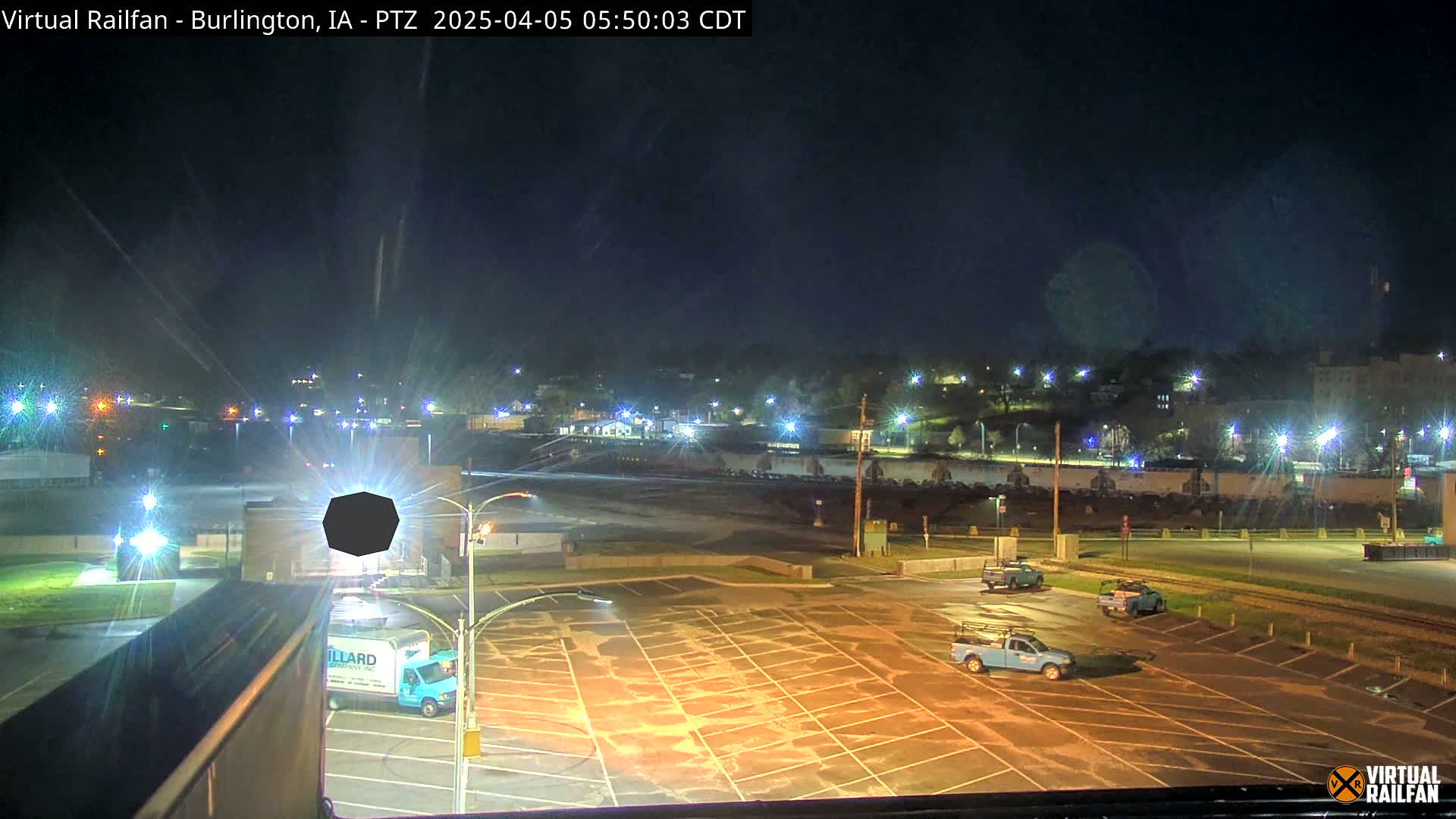 A nighttime view of a mostly empty parking lot with a few trucks, and a city illuminated by streetlights in the background under a clear sky.
