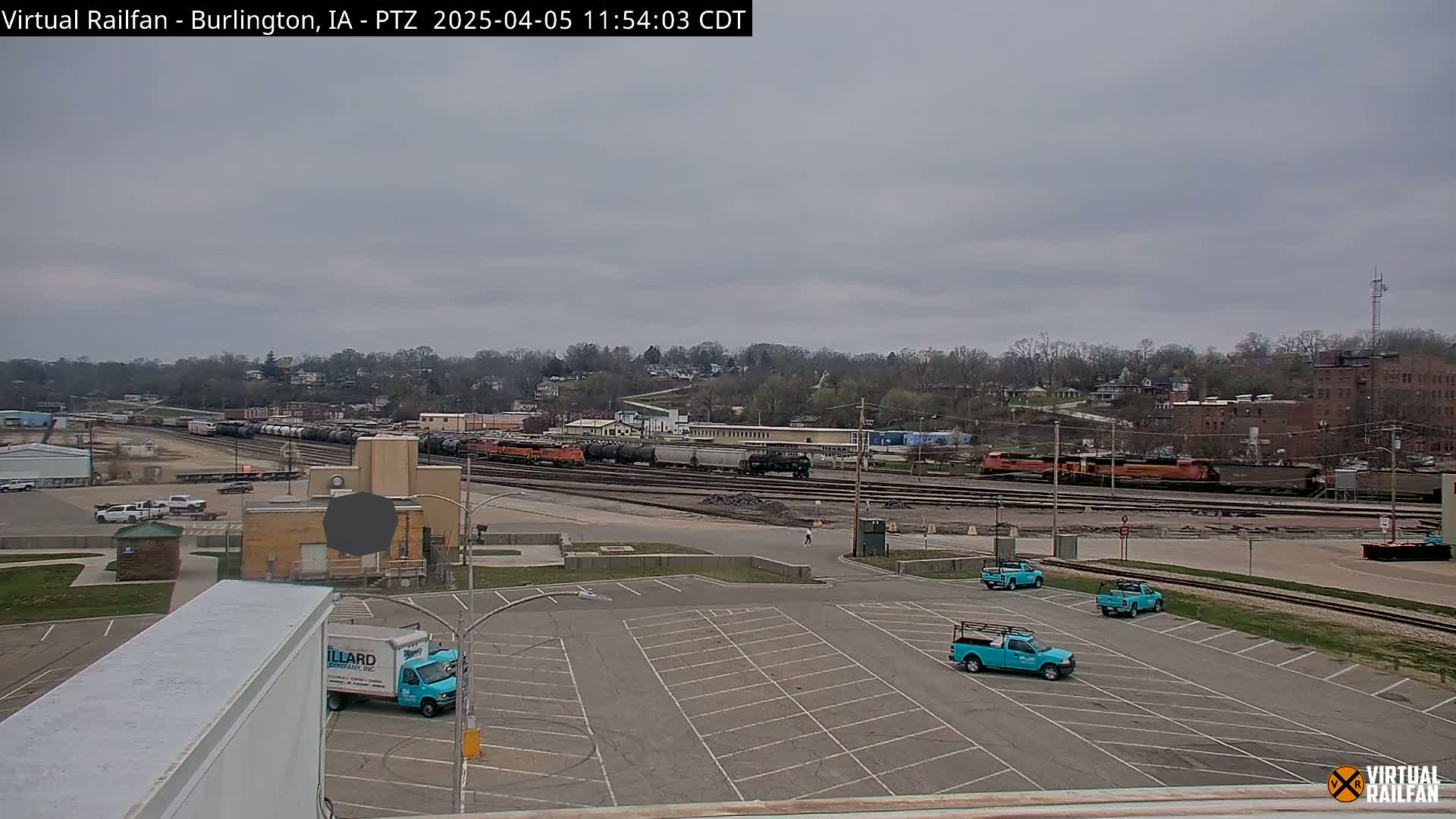 A mostly cloudy day reveals a railroad yard with several trains and a parking lot containing several teal pickup trucks.