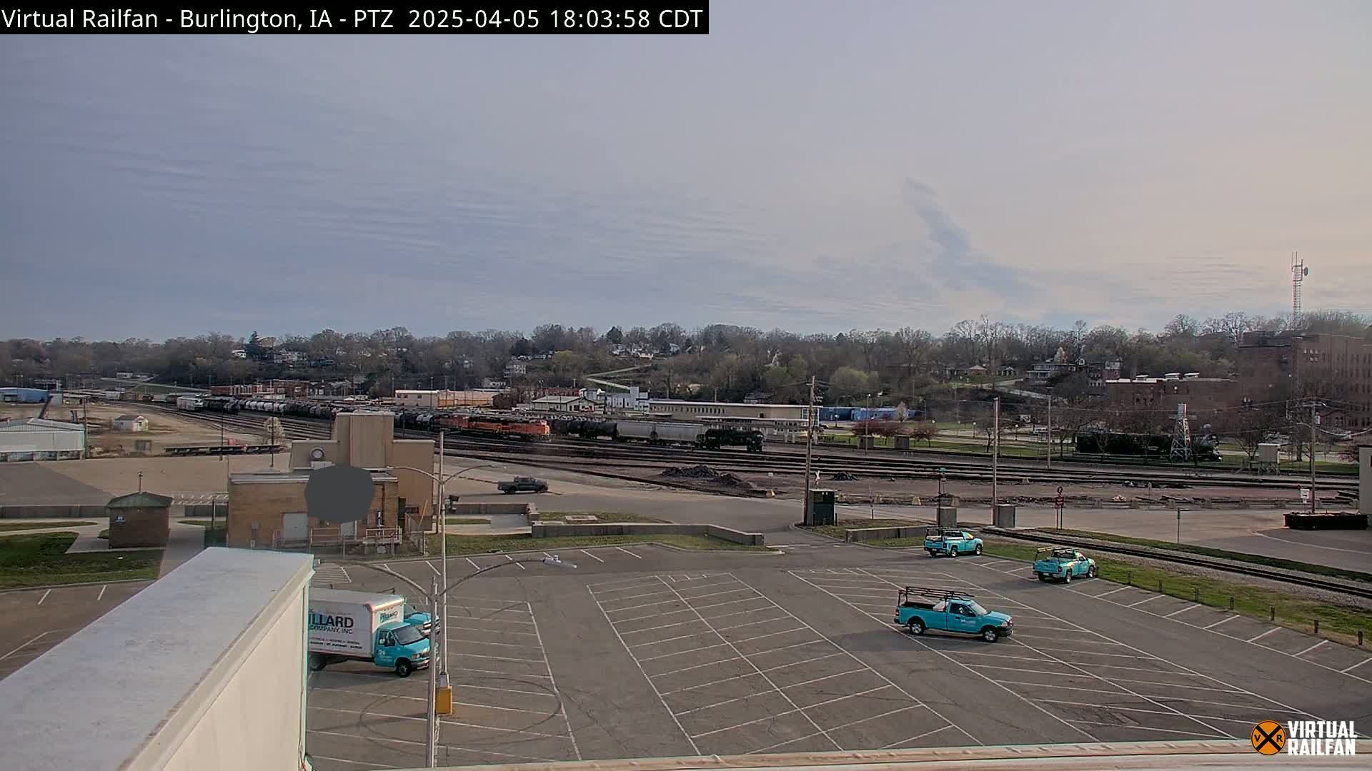 A mostly empty parking lot with several teal pickup trucks is in the foreground, overlooking a railroad yard with numerous train cars and a tree-lined hillside under a mostly clear, light sky.
