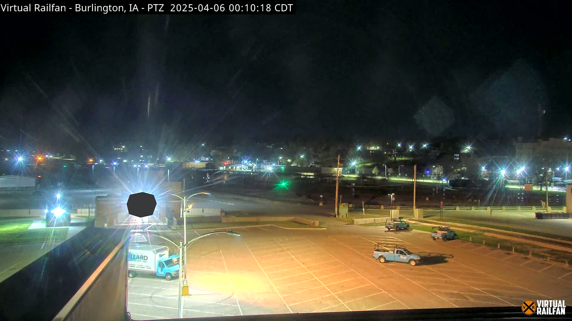 A mostly empty parking lot at night, illuminated by streetlights, with a few trucks parked and a distant town visible under a clear, dark sky.