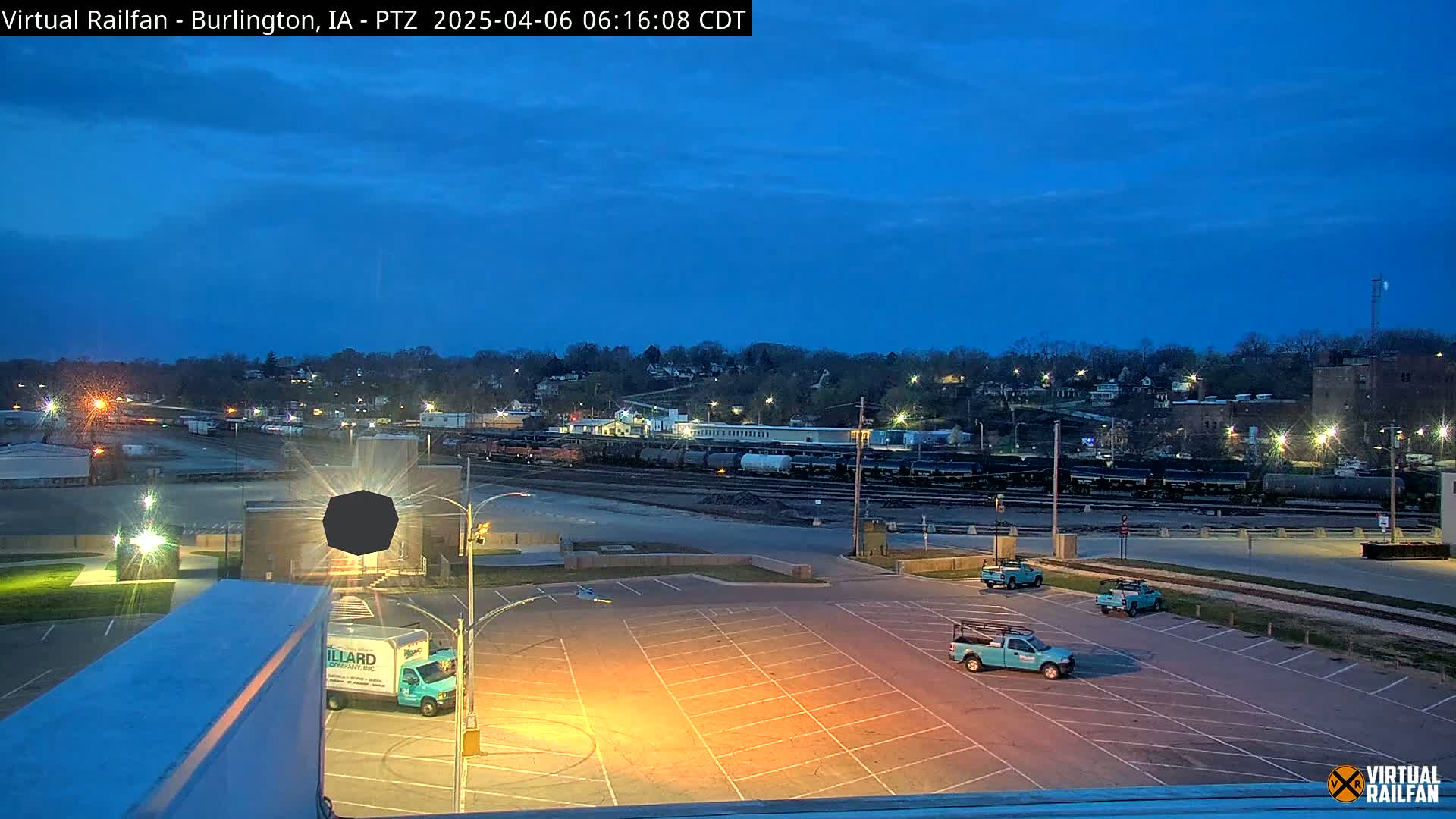 Under a twilight blue sky, a mostly empty parking lot sits in front of a rail yard with several parked trucks, and a hill with buildings and trees in the background.