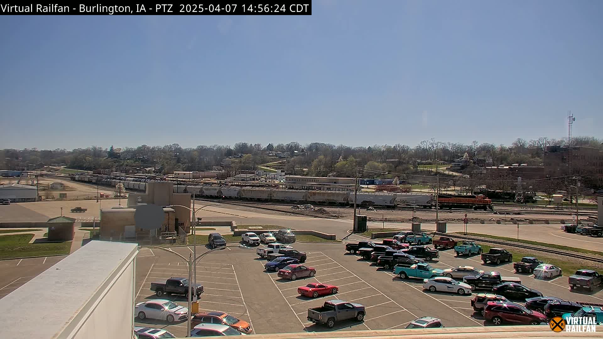 A mostly sunny day reveals a train yard with numerous railcars and a locomotive, along with a parking lot filled with various cars, all situated near a tree-lined hillside.