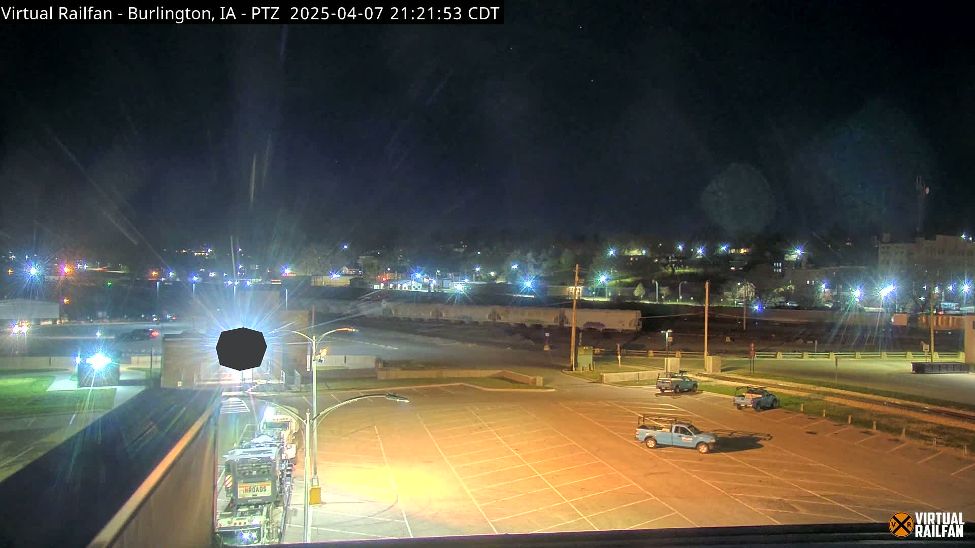 A nighttime view of a mostly empty parking lot with several work trucks, a train in the background, and a cityscape illuminated by streetlights under a clear, dark sky.