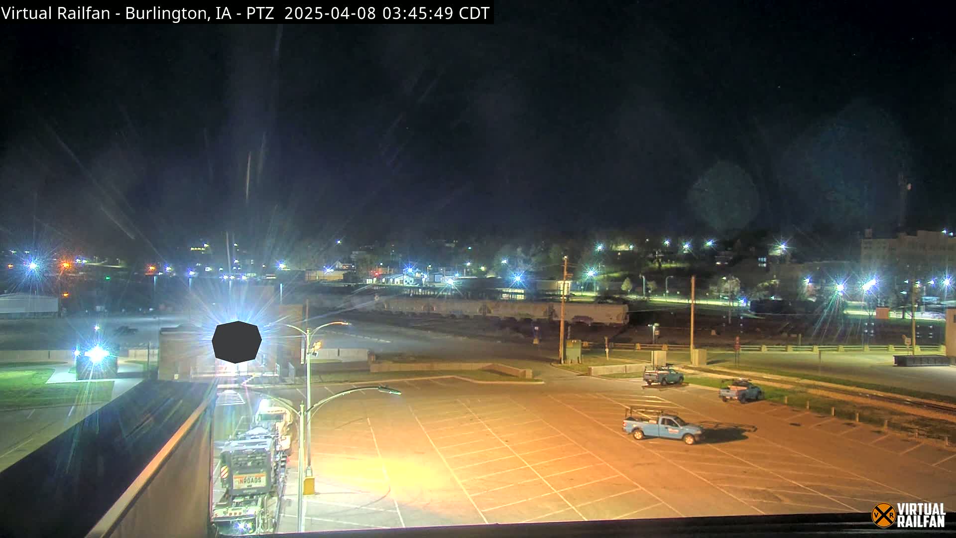 A nighttime view of a mostly empty parking lot next to railroad tracks, with several vehicles parked and a few buildings visible in the distance under a clear sky.