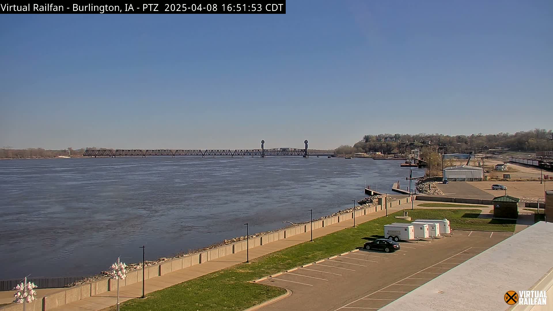 A wide river with a railroad bridge in the distance, under a clear blue sky, is viewed from an elevated vantage point overlooking a paved walkway and a parking lot.