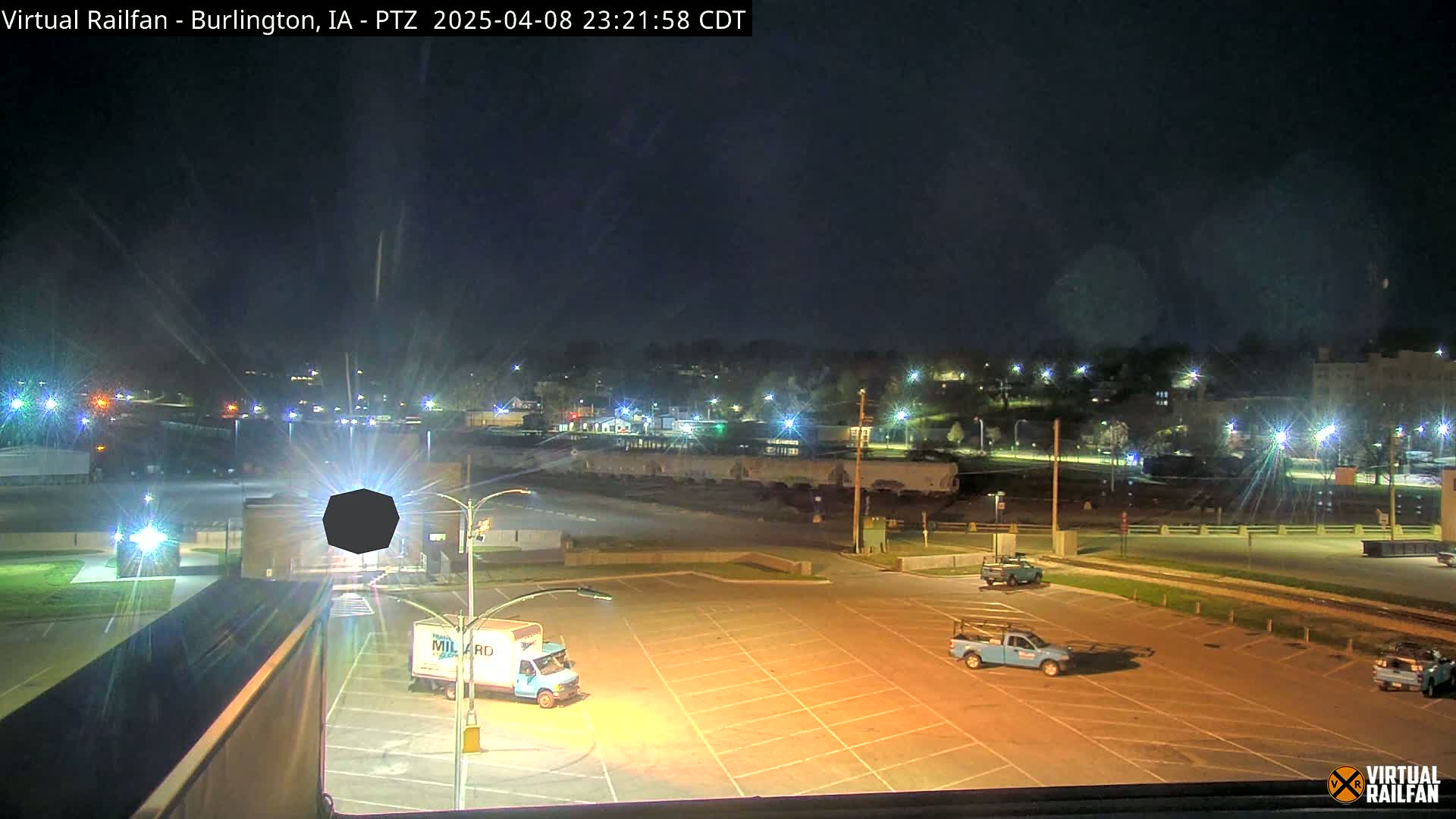 A mostly empty, brightly lit parking lot at night, with two trucks parked, and a distant town illuminated by streetlights under a dark sky.