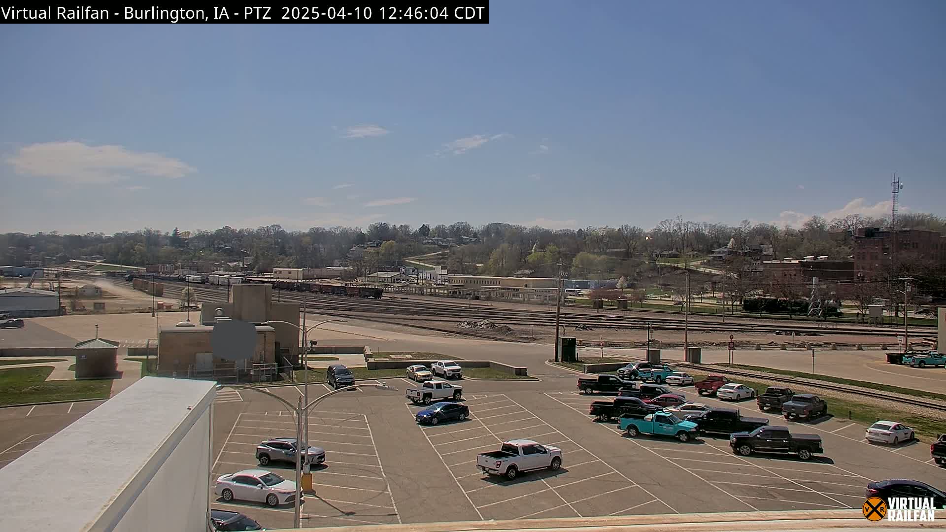 A mostly sunny day reveals a train yard with several cars parked in a lot in the foreground, and a line of trees and buildings in the background.