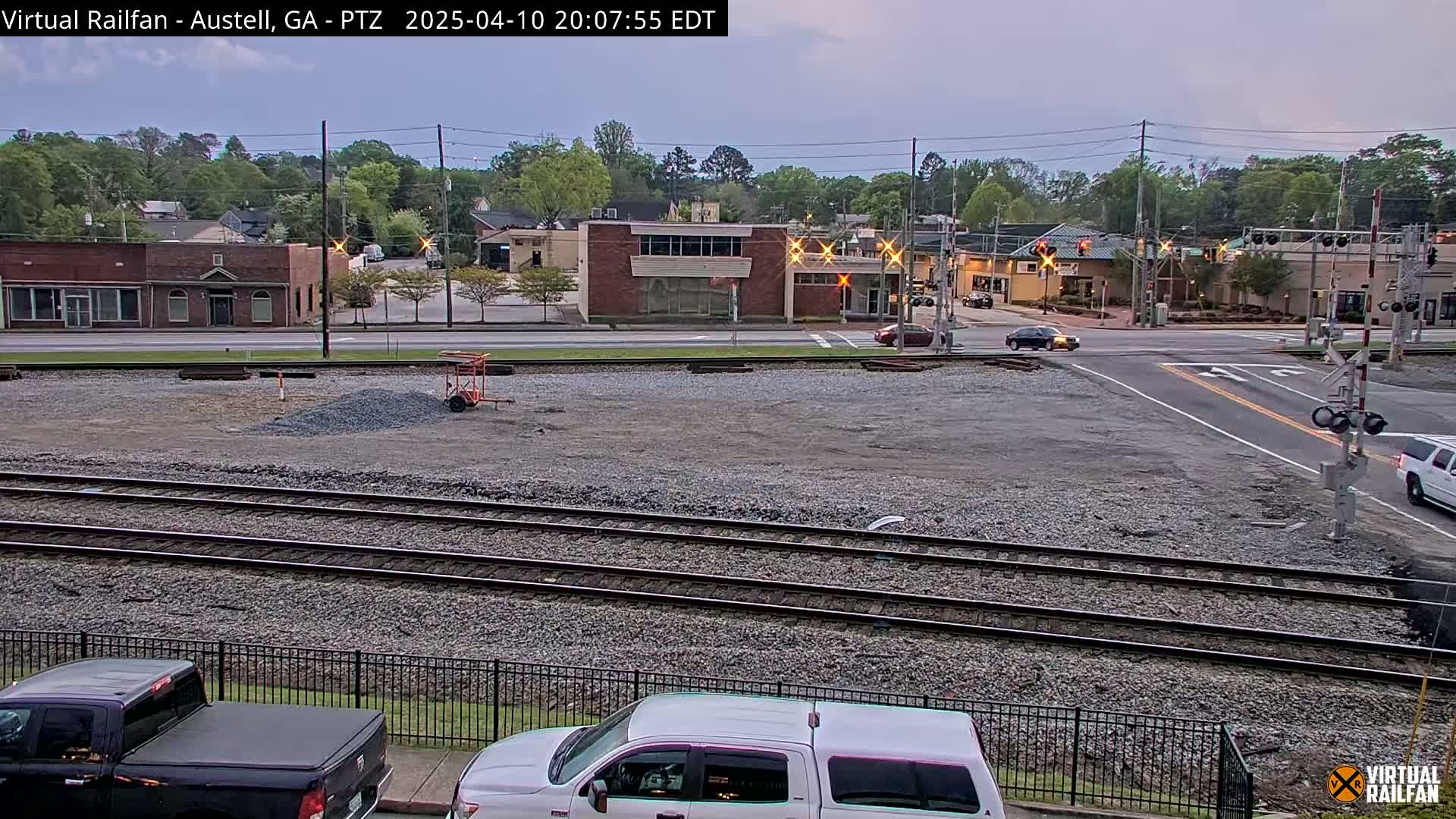 Under an overcast sky, a view of railroad tracks, a gravel area, and a street intersection shows several buildings and cars in the background.