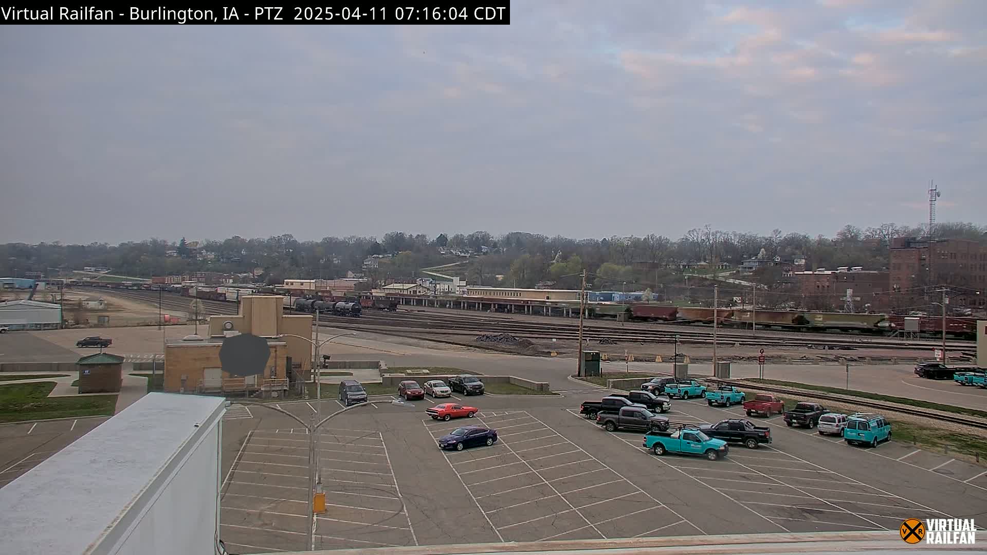 An overcast day shows a railroad yard with multiple train cars and a parking lot filled with several cars and trucks.