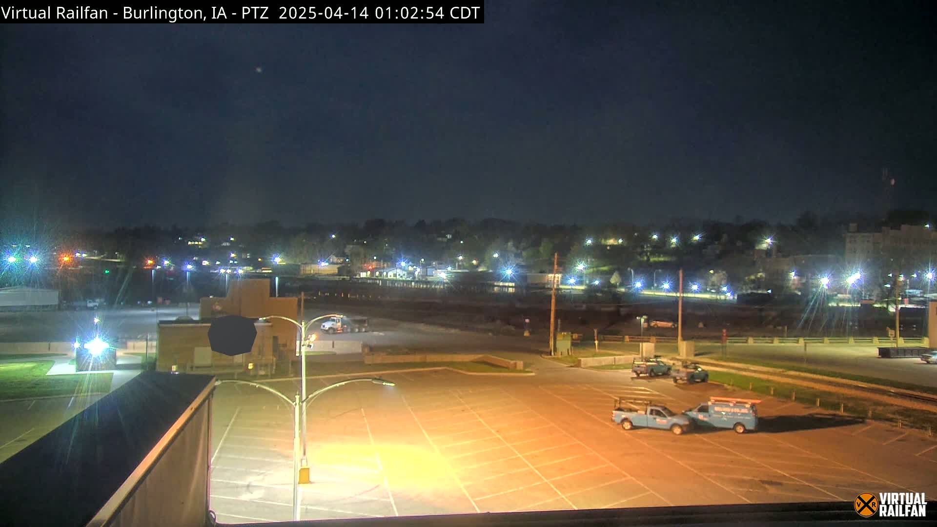 A nighttime view of a mostly empty parking lot with a few parked vehicles, overlooking a town illuminated by streetlights under a clear, dark sky.