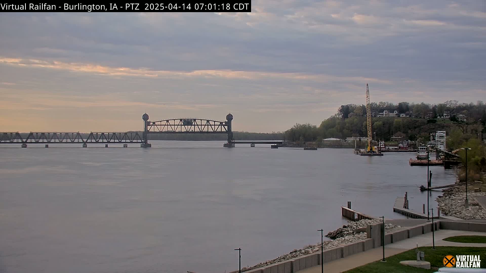 A river scene under a cloudy sky shows a metal bridge, a construction crane on a barge, and a shoreline with a paved walkway.