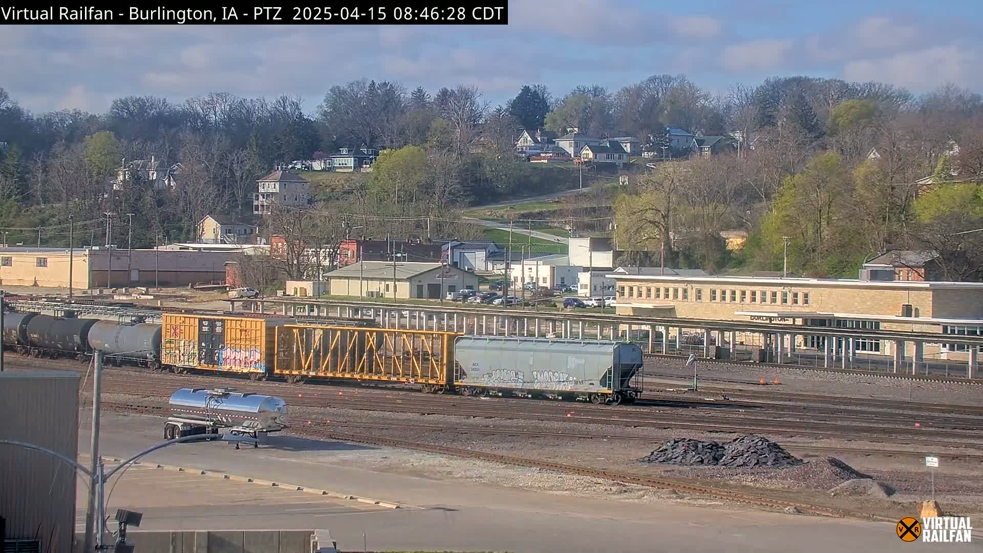 A train with several cars sits on railroad tracks in front of a long building, with a hillside of trees and houses visible in the background under a partly sunny sky.