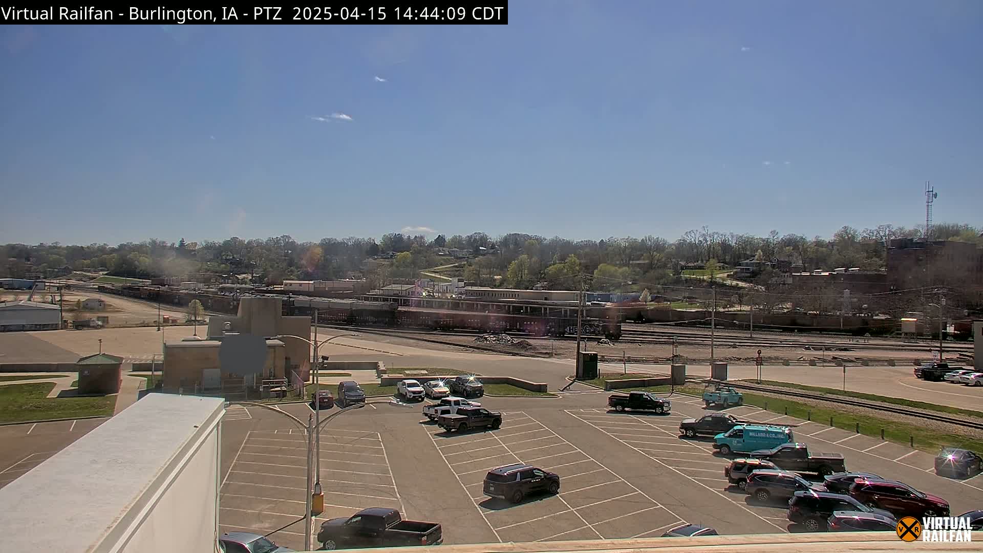 A mostly sunny day reveals a train yard with several rail cars and a parking lot with numerous vehicles, all situated in front of a tree-lined hill.
