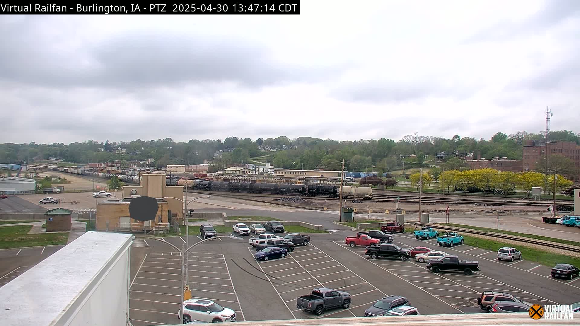 A mostly cloudy day reveals a train yard with many rail cars, a parking lot filled with vehicles, and a hilly, tree-covered background.