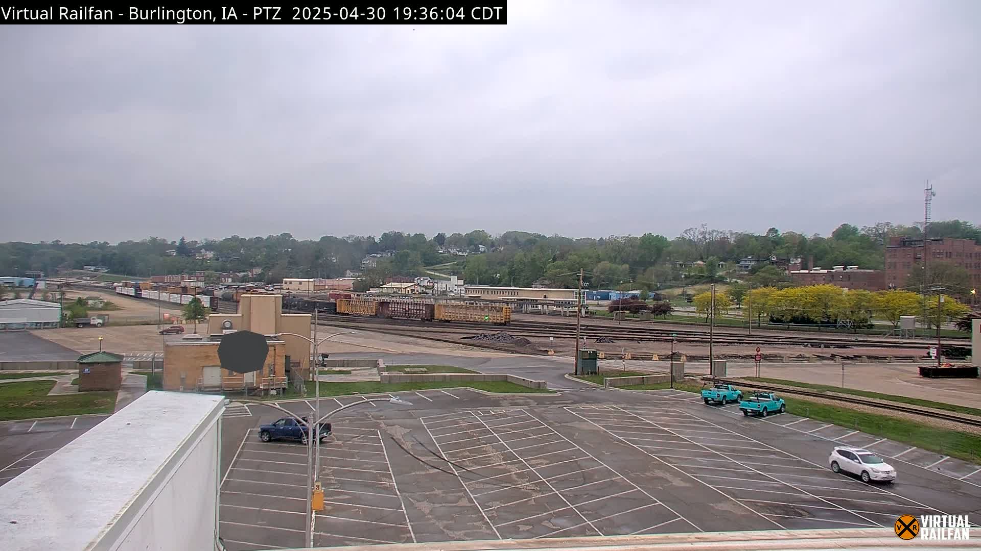 A mostly empty parking lot overlooks a railroad yard with several train cars and a few vehicles, all under an overcast sky.