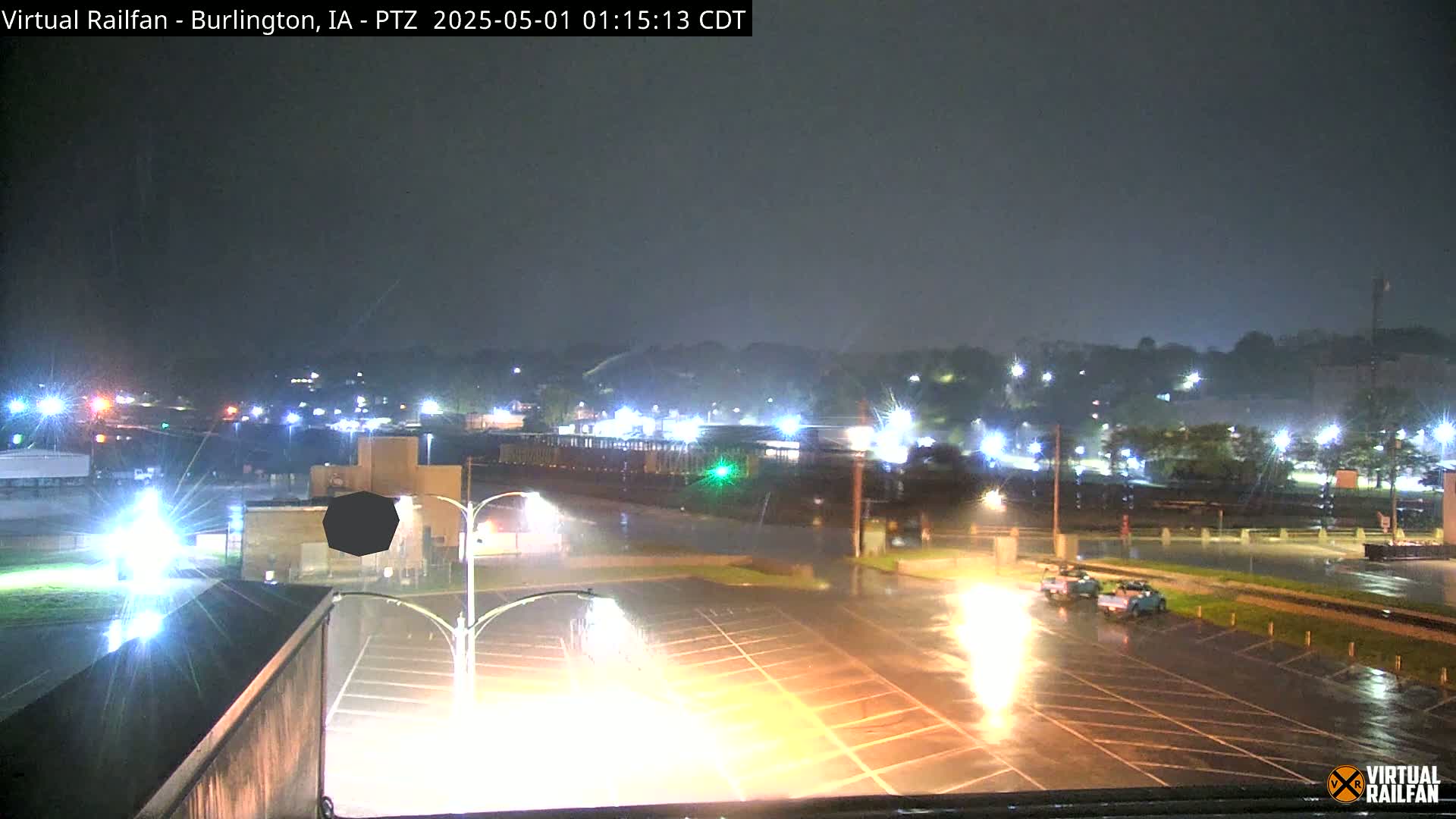 A nighttime view of a rain-soaked parking lot and surrounding area, illuminated by streetlights, with vehicles visible on a wet road.