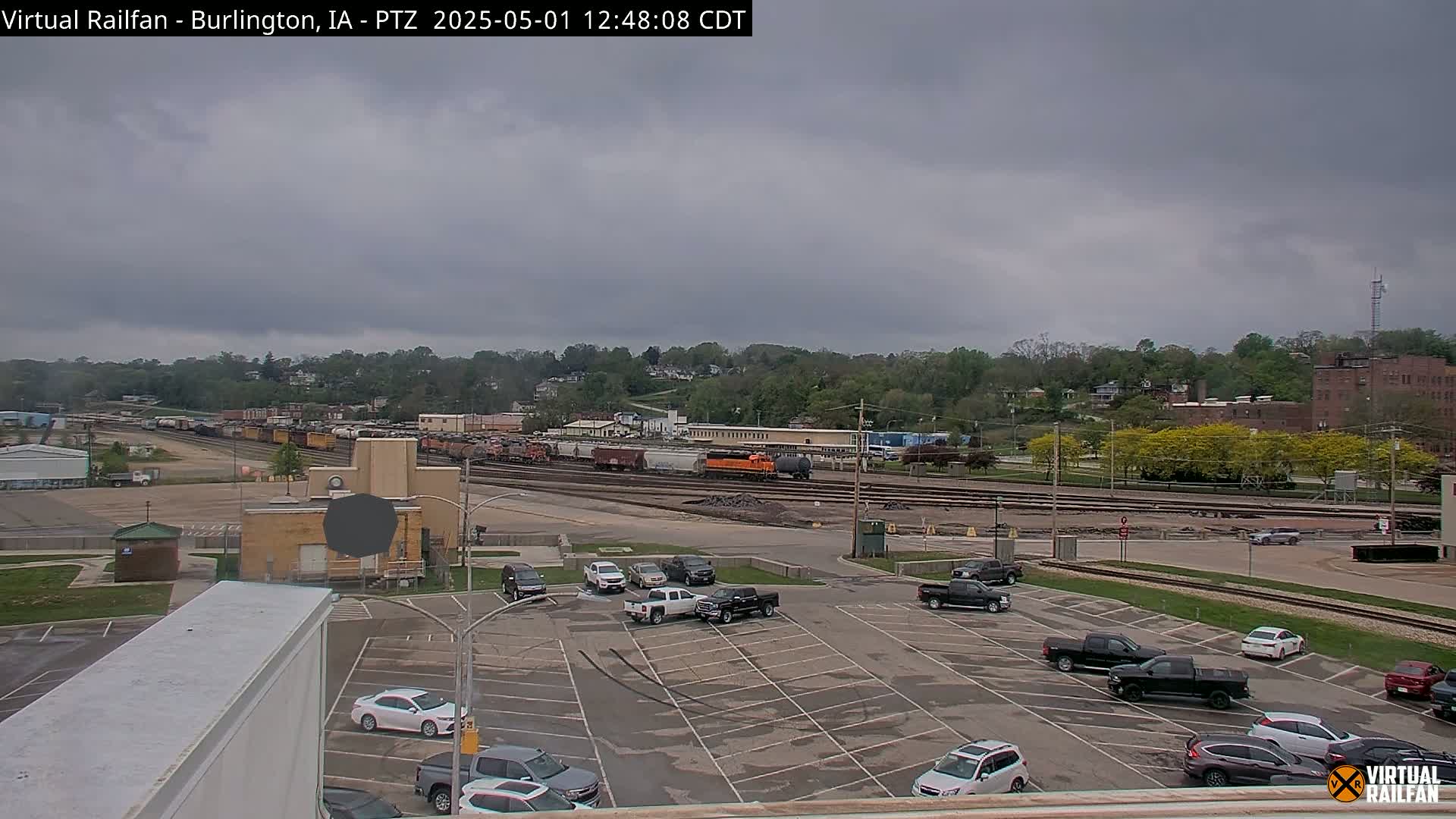 A mostly cloudy day reveals a view of a large rail yard with numerous train cars, a parking lot with several vehicles, and a background of trees and buildings.