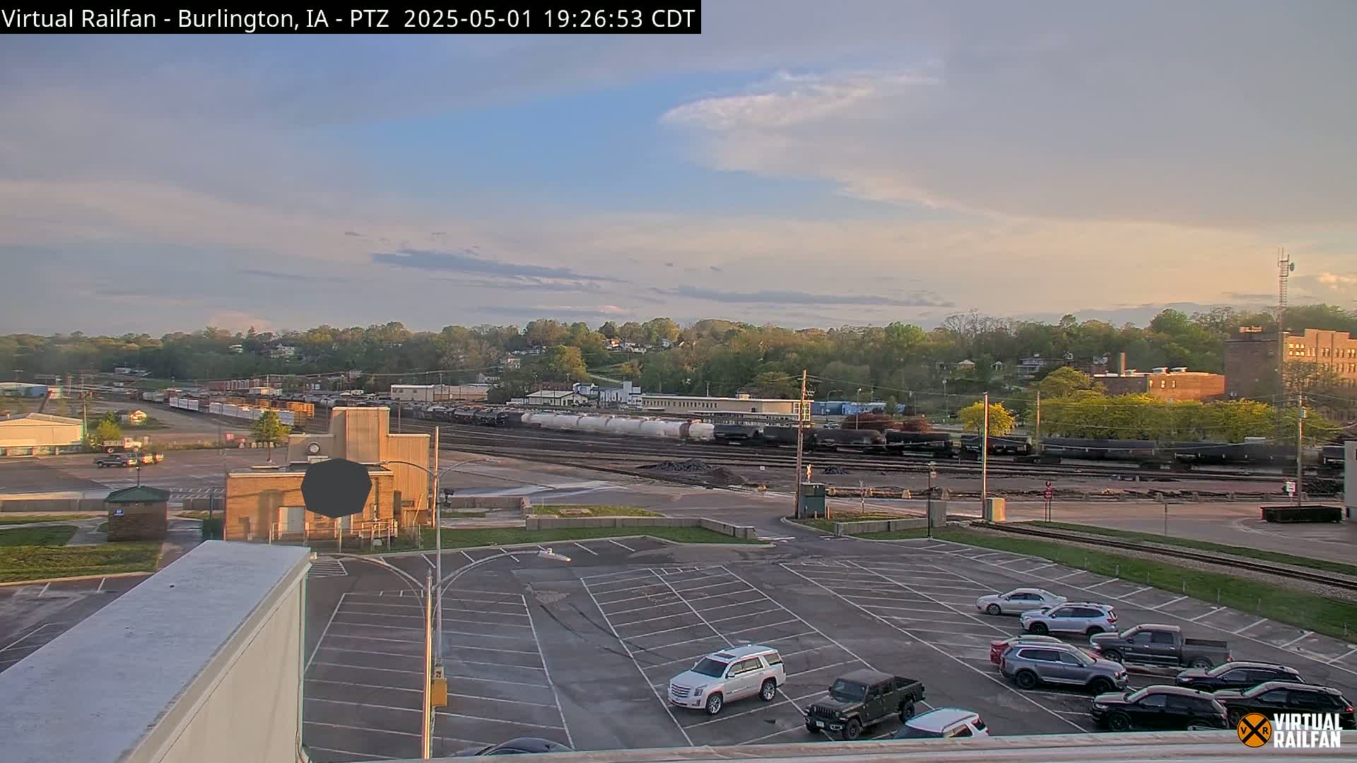 A mostly sunny evening scene shows a large parking lot with several cars, adjacent to a rail yard filled with train cars, and a tree-lined hill in the background.