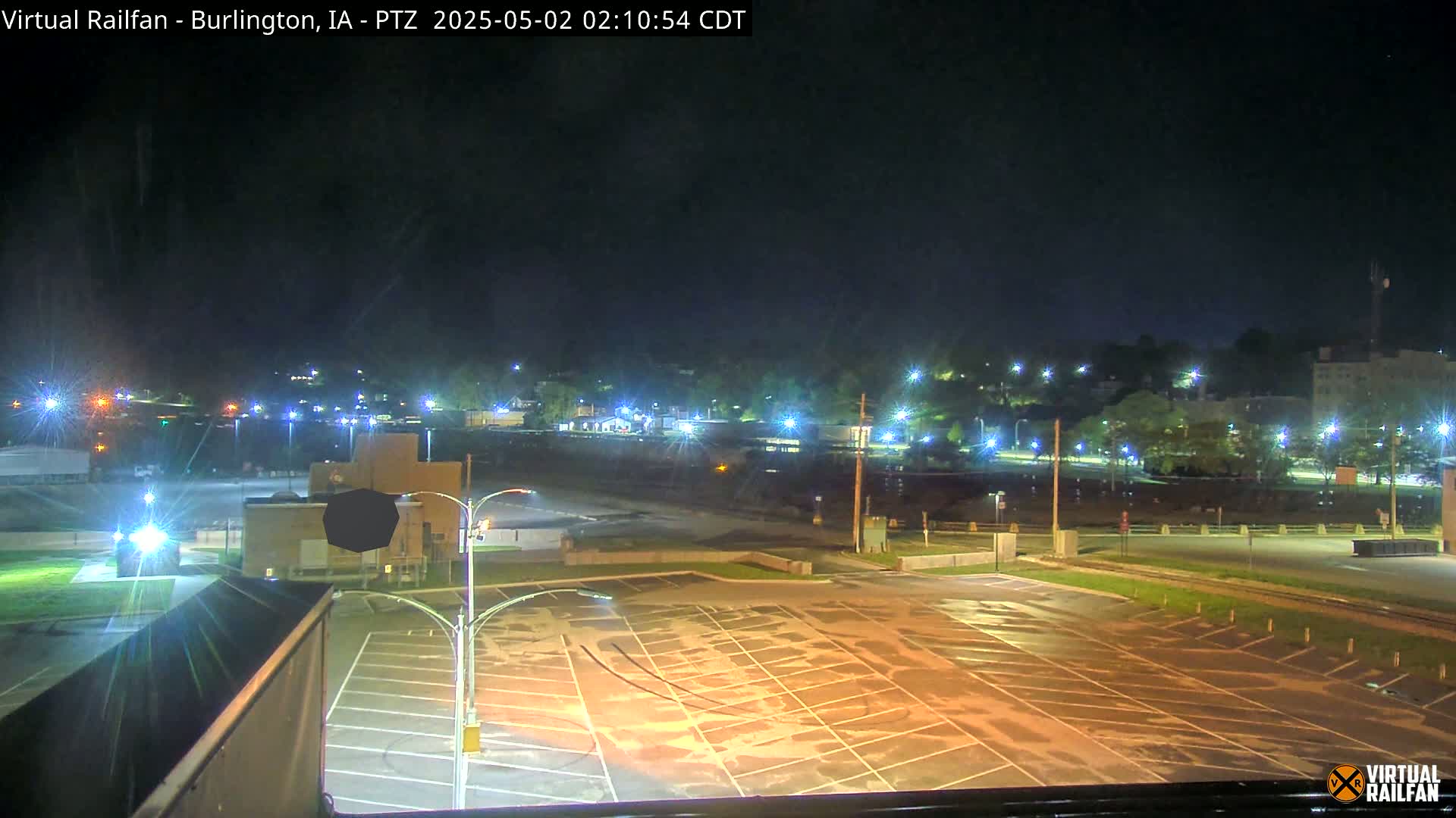 A nighttime view of a mostly empty parking lot illuminated by lights, with a dimly lit cityscape and trees in the background under a clear sky.