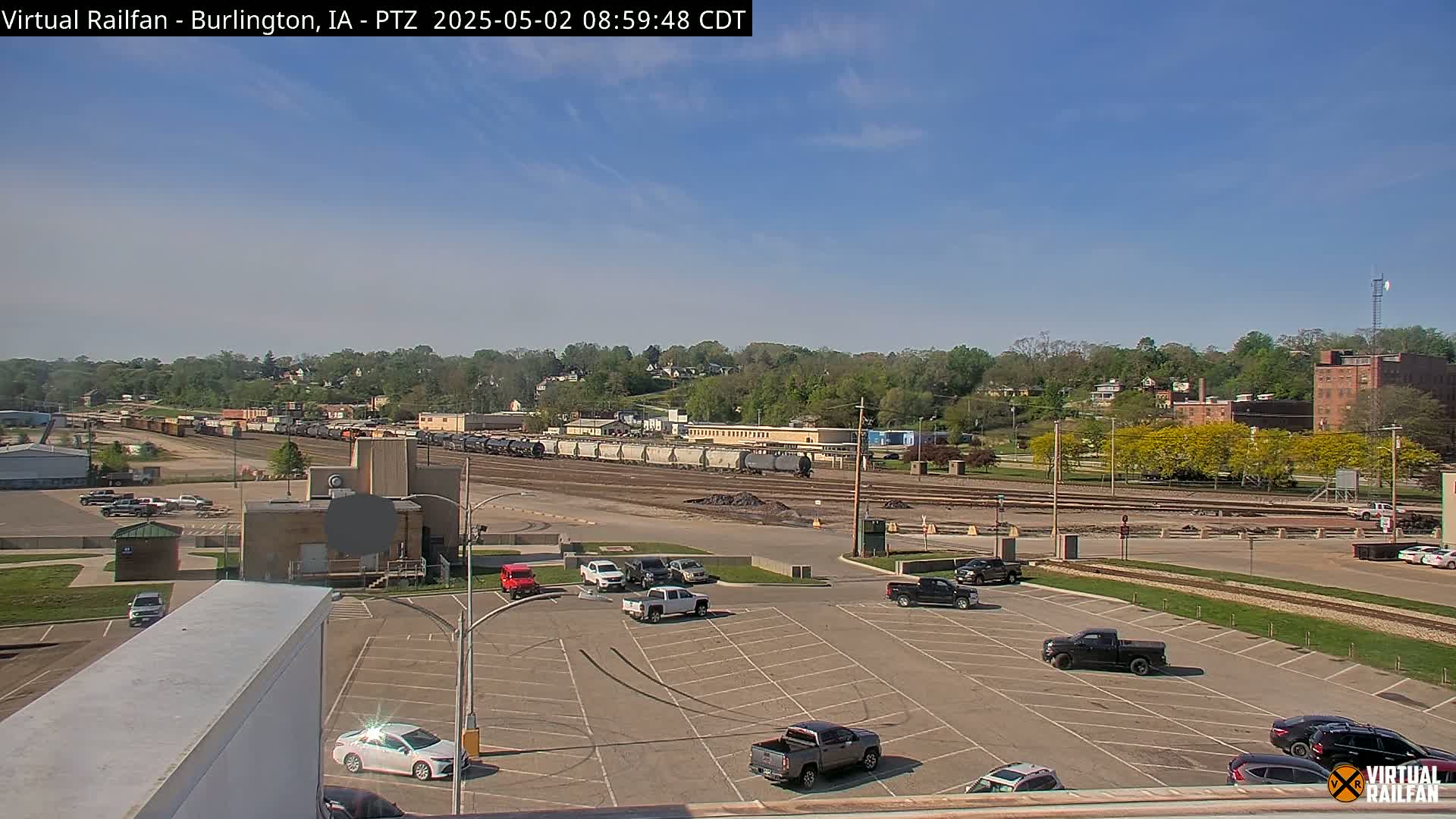 A mostly sunny day reveals a train yard with multiple rail cars, a parking lot with several vehicles, and a tree-covered hillside in the background.