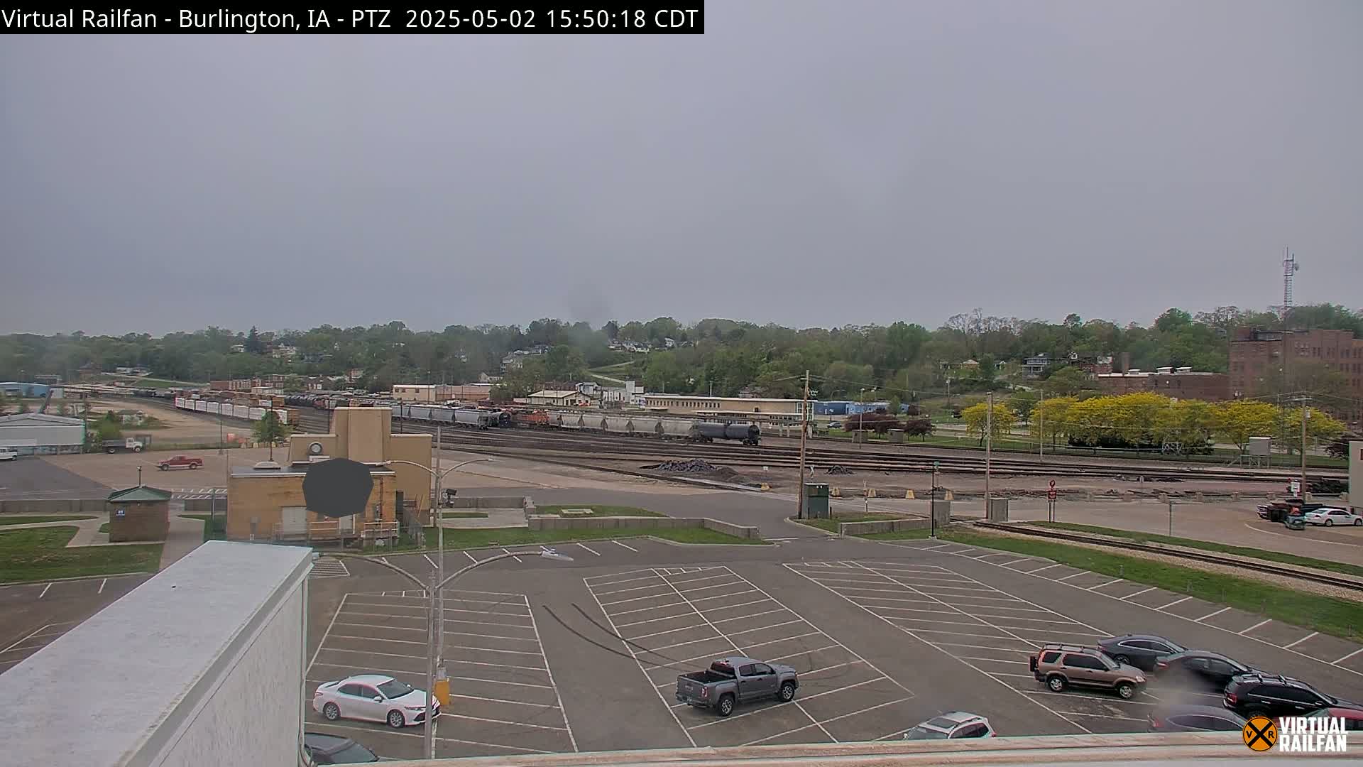 An overcast day reveals a train yard with several railcars, a parking lot with a few vehicles, and a tree-lined hillside in the background.