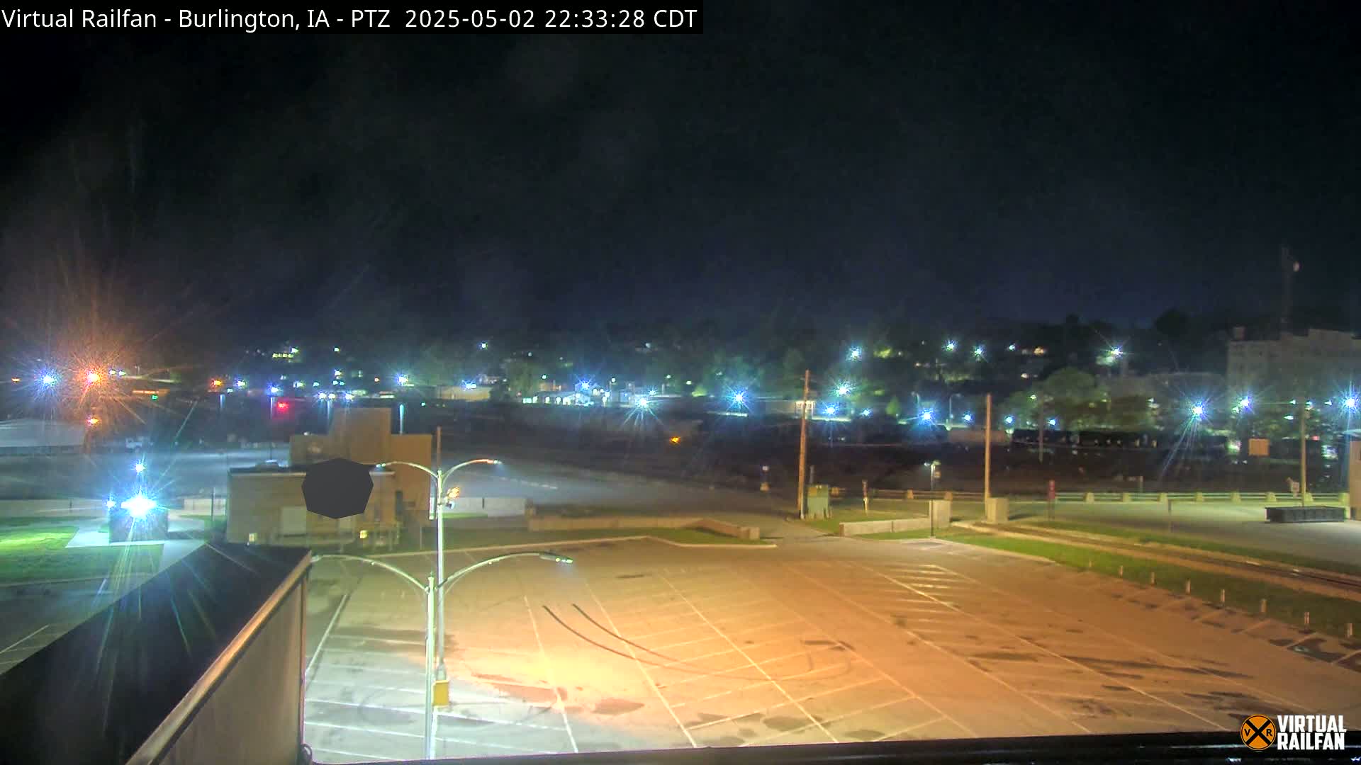 A nighttime view of an empty parking lot illuminated by streetlights, with a distant cityscape and train tracks visible under a clear sky.