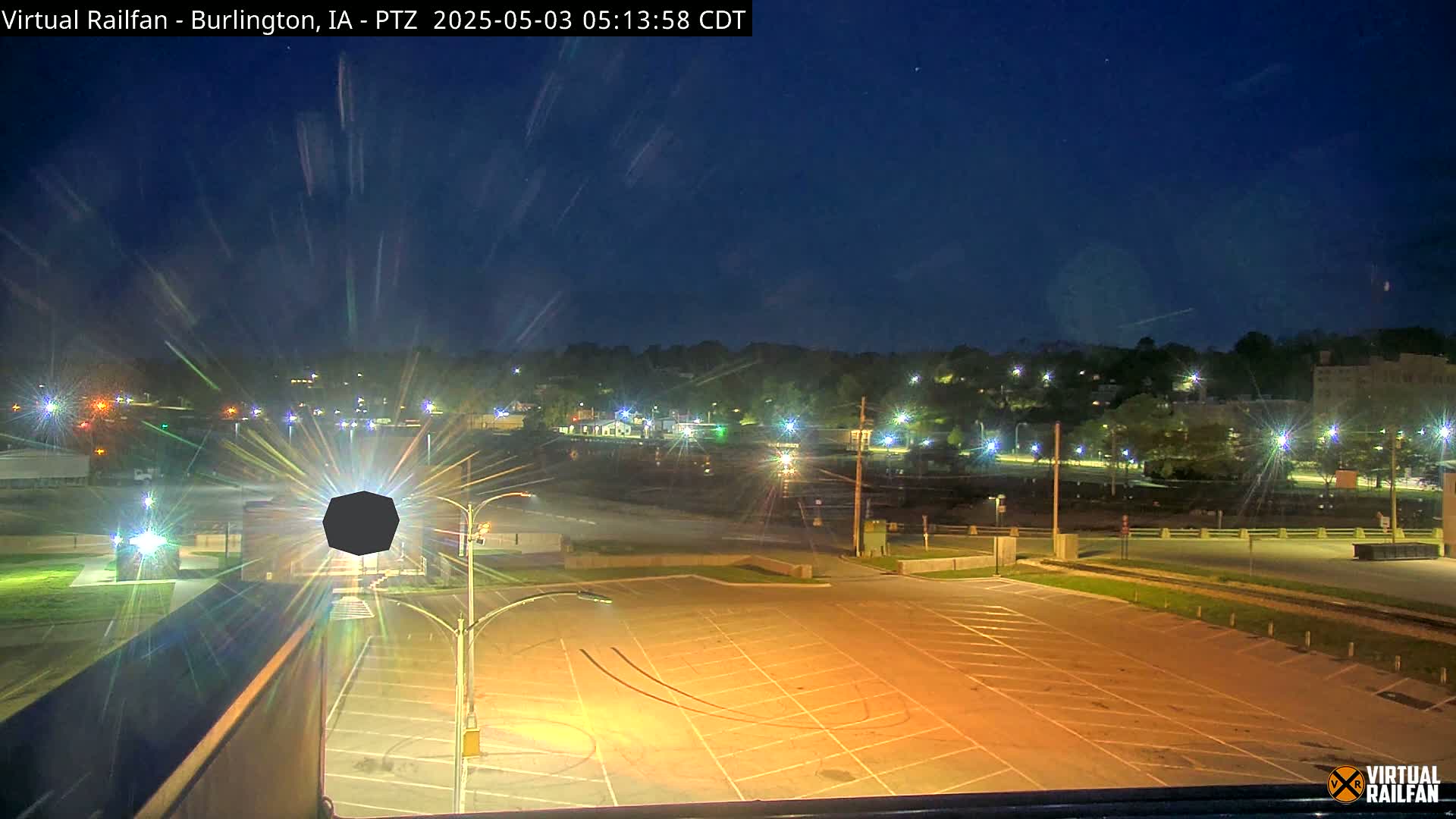 An empty, illuminated parking lot overlooks a dimly lit town at night under a dark blue, clear sky.