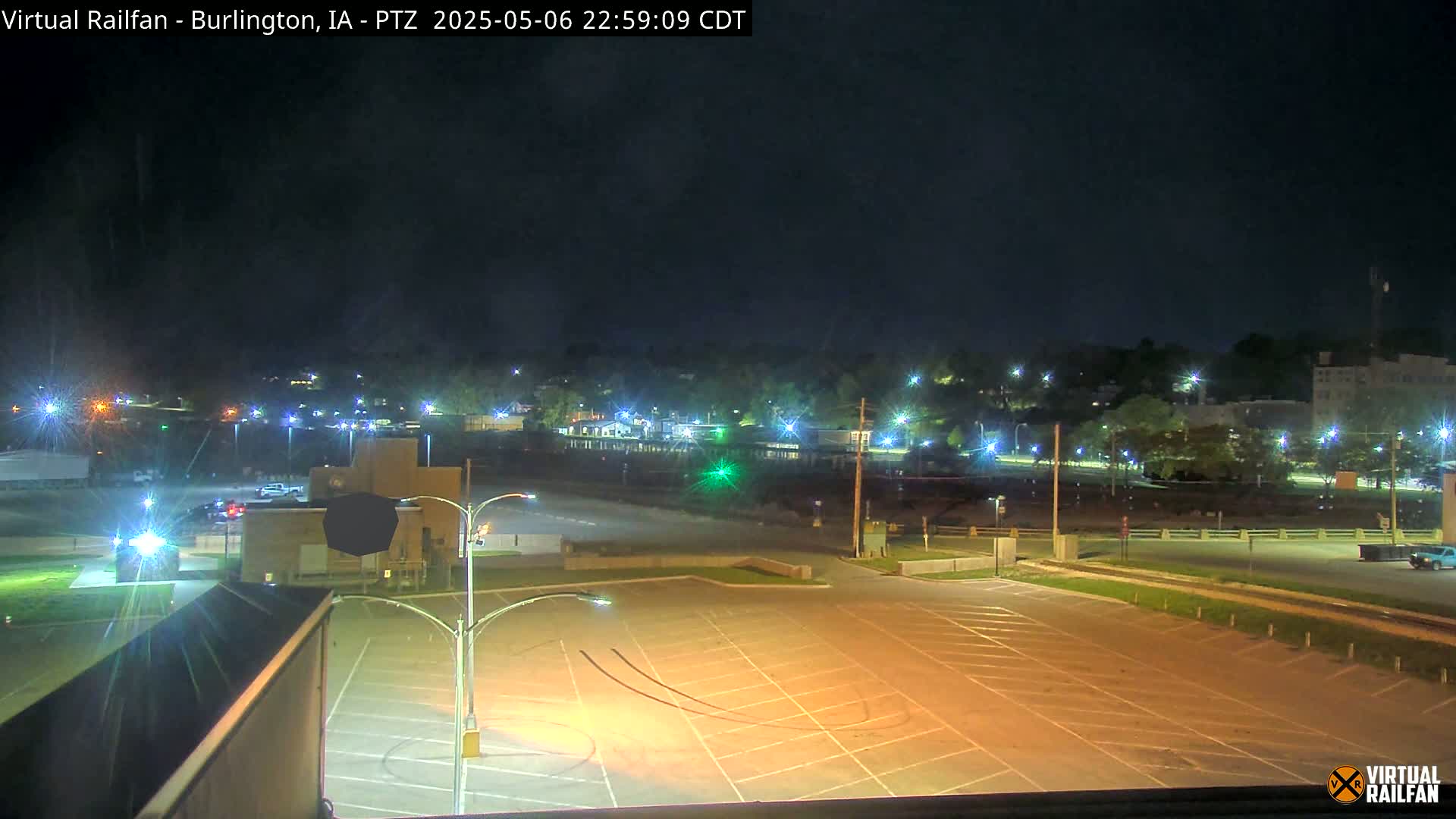 A mostly empty, illuminated parking lot at night overlooks a dimly lit town across a street and railroad tracks.