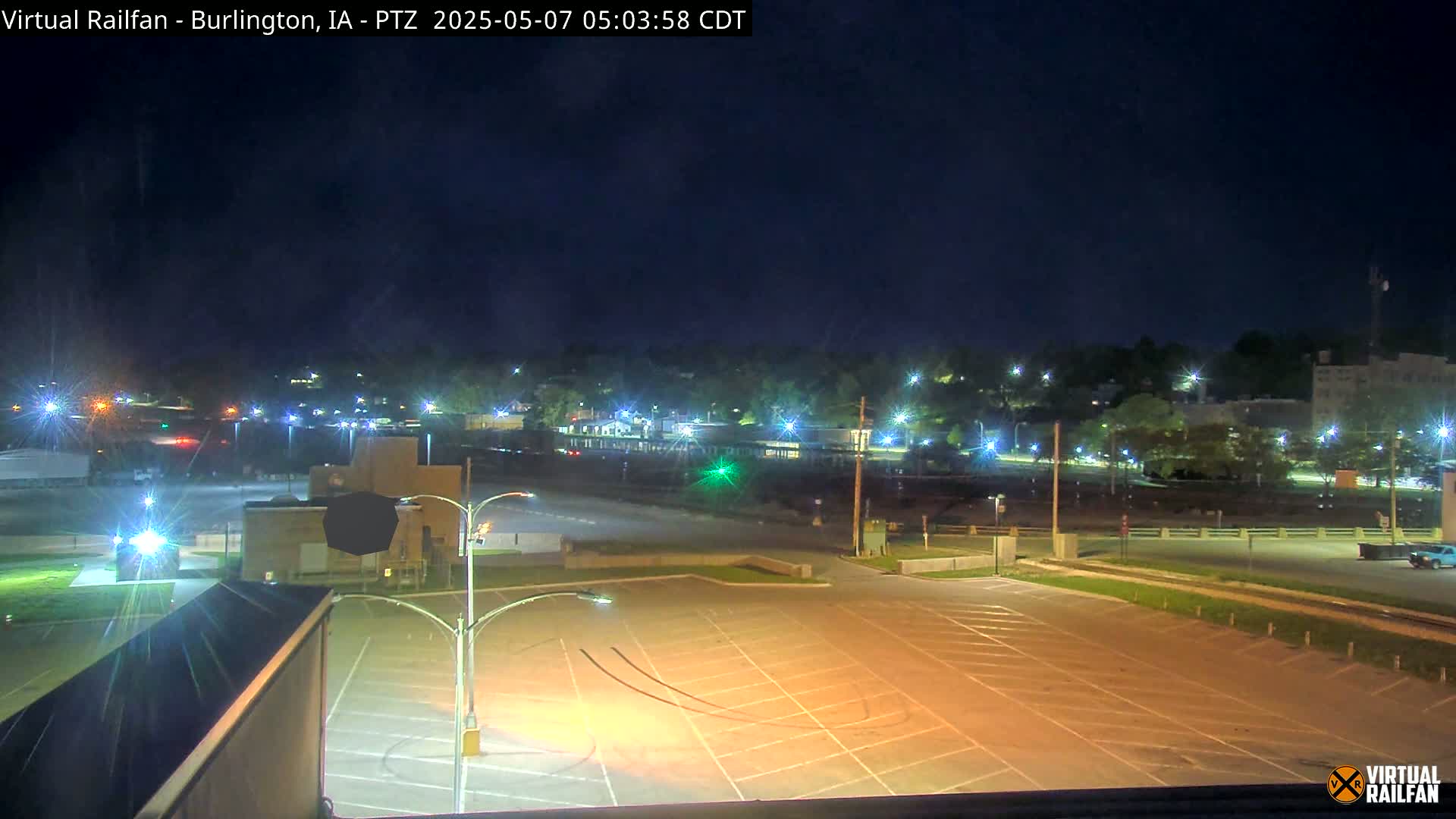 An empty parking lot at night is illuminated by streetlights, overlooking a dimly lit town under a dark, clear sky.