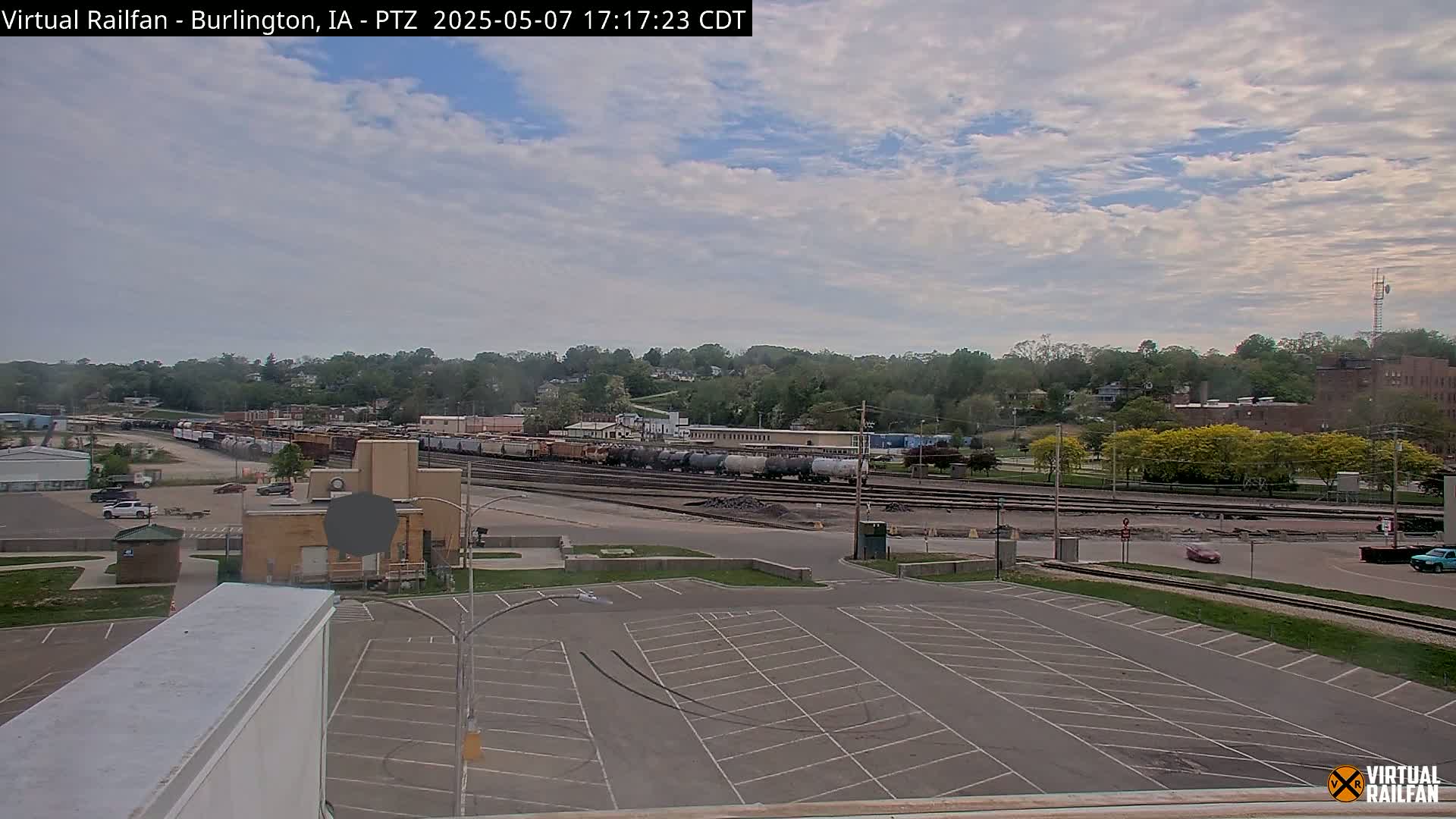 A partly cloudy sky overlooks a train yard with several railcars, a parking lot, and a tree-covered hill in the background.
