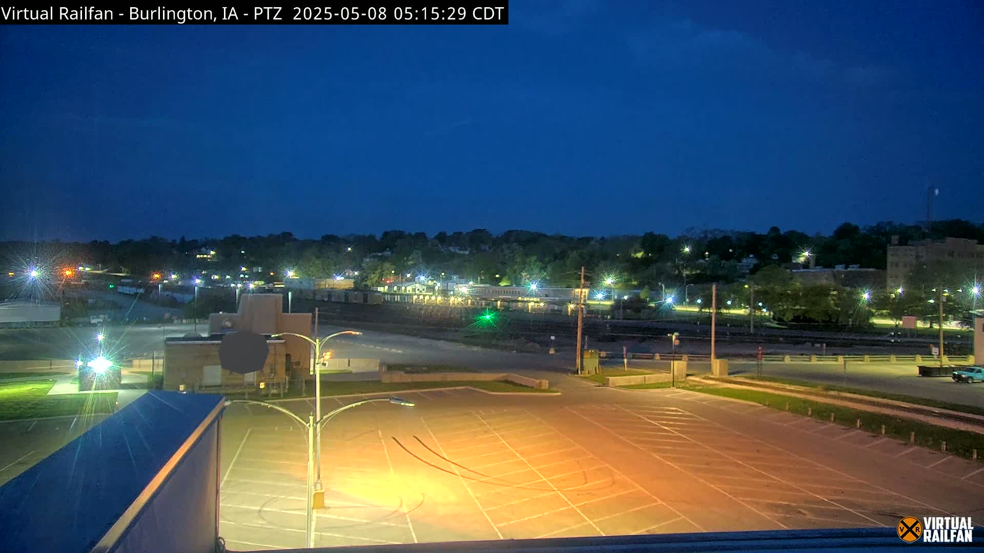 An empty, illuminated parking lot overlooks a nighttime cityscape under a dark blue, clear sky.