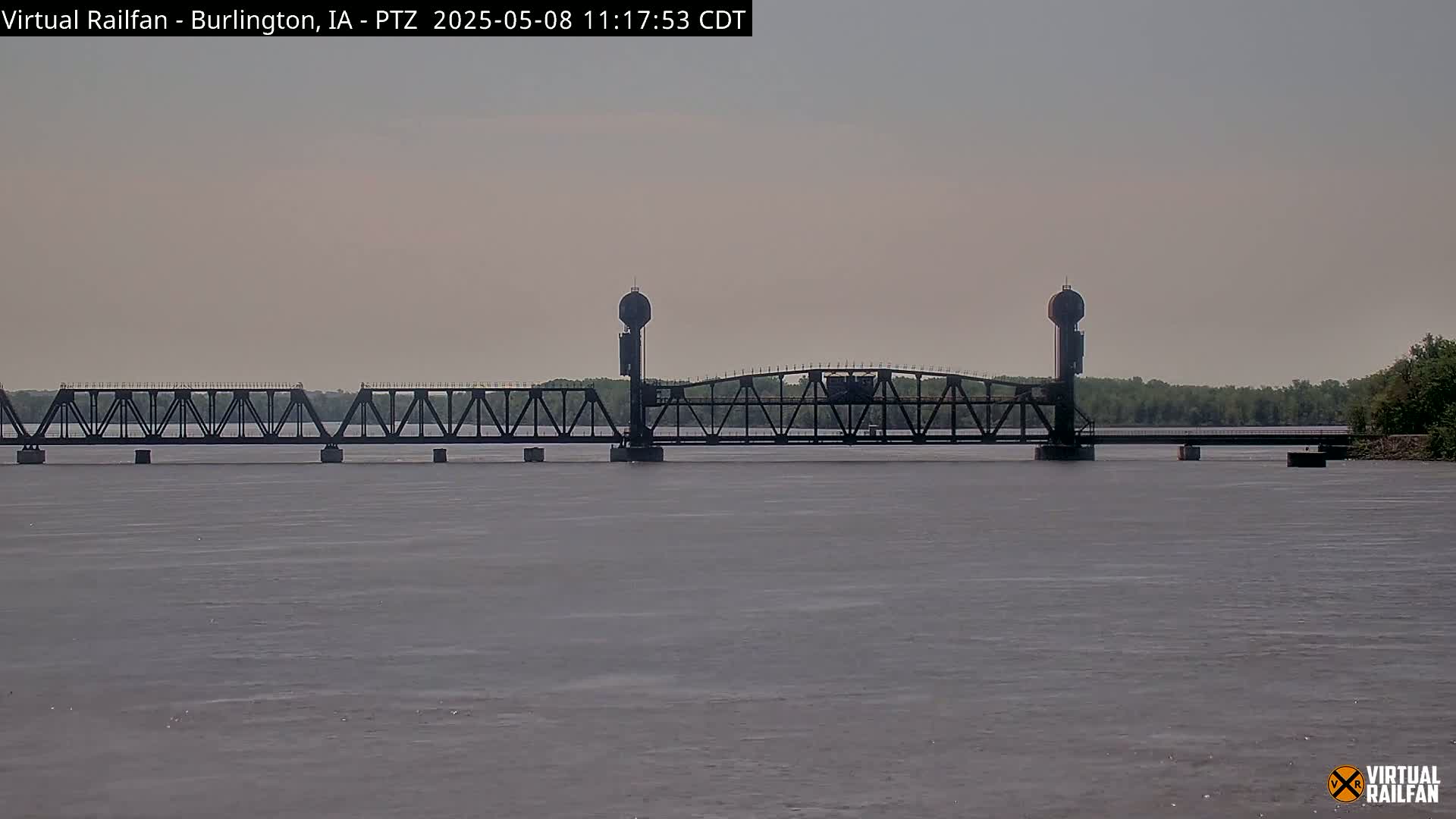 A long, dark metal bridge spans a calm, gray river under a hazy sky.