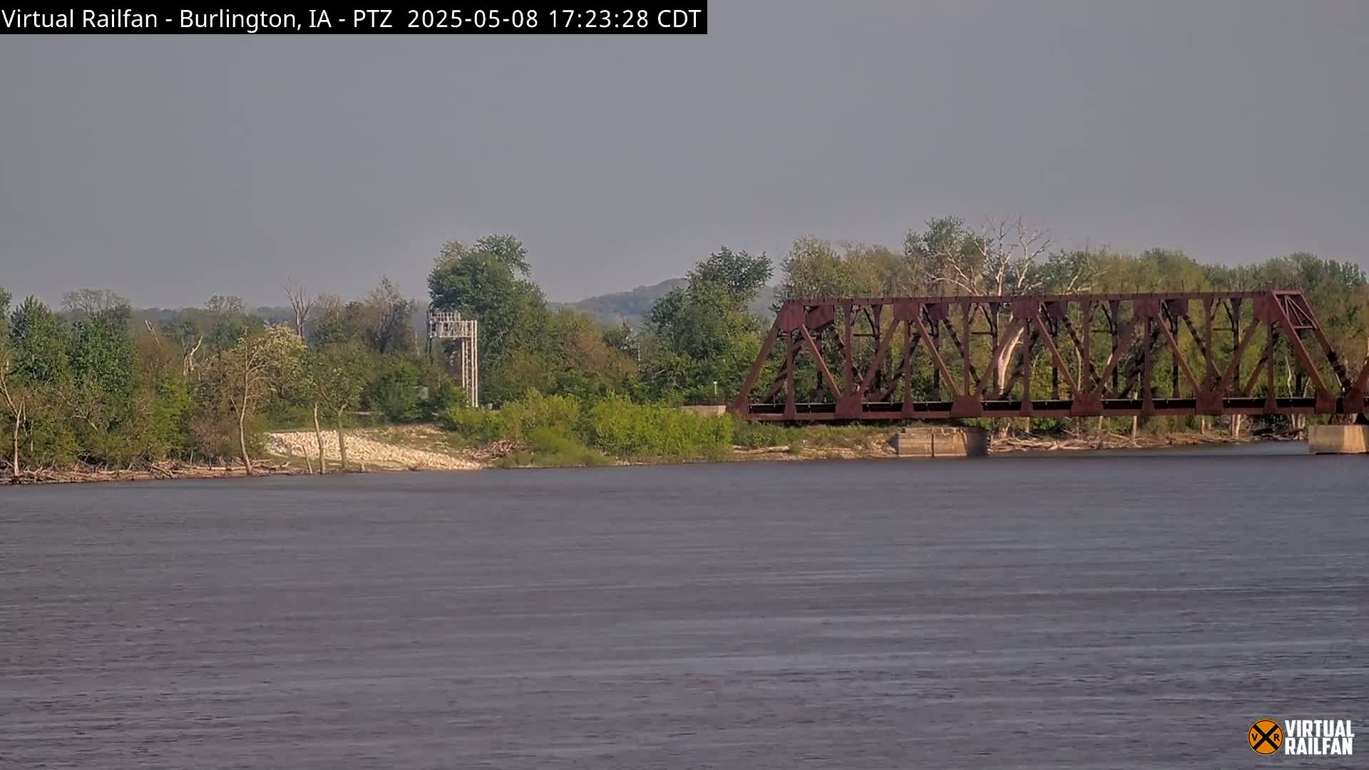 A rusty railroad bridge spans a wide, calm river, with green trees and a tall structure on the opposite bank under a hazy sky.
