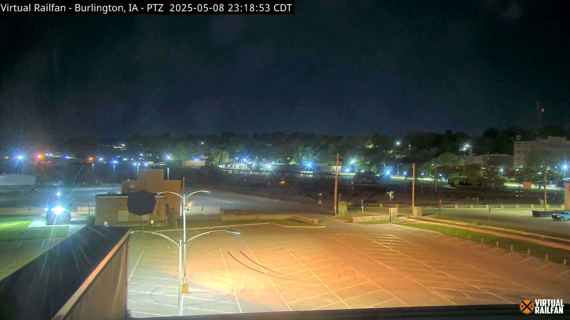 An empty, illuminated parking lot at night overlooks a town with streetlights visible under a clear, dark sky.