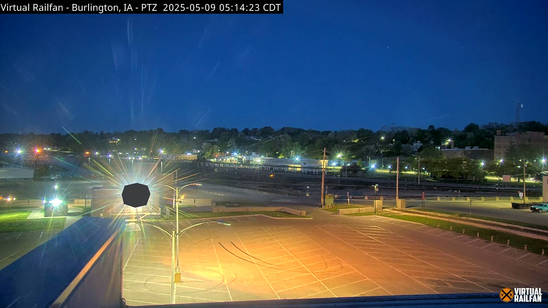 An empty, illuminated parking lot at twilight is situated in front of a dark, tree-lined hill with distant buildings and lights.