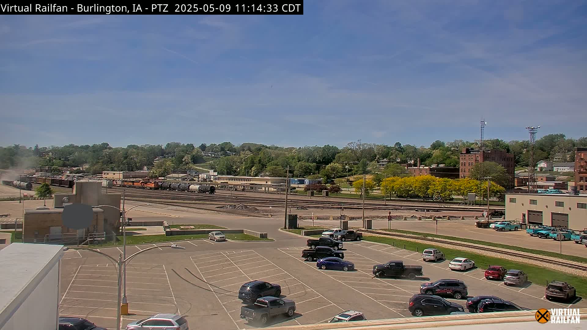 A partly sunny day shows a train yard with numerous railcars, a parking lot with several cars, and a hillside covered in green trees and some buildings in the background.